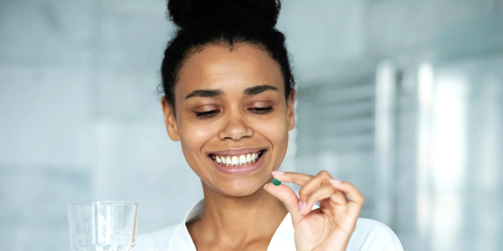 A woman smiling and holding a green pill between her thumb and index finger in a bright, modern setting.