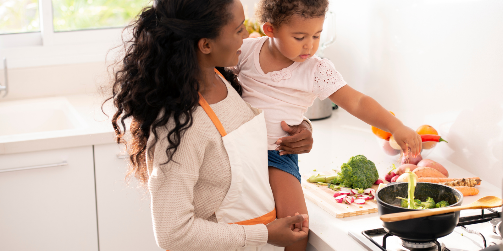 A woman and a young girl cooking together in a bright kitchen, with vegetables like radishes, broccoli, and carrots on the counter.
