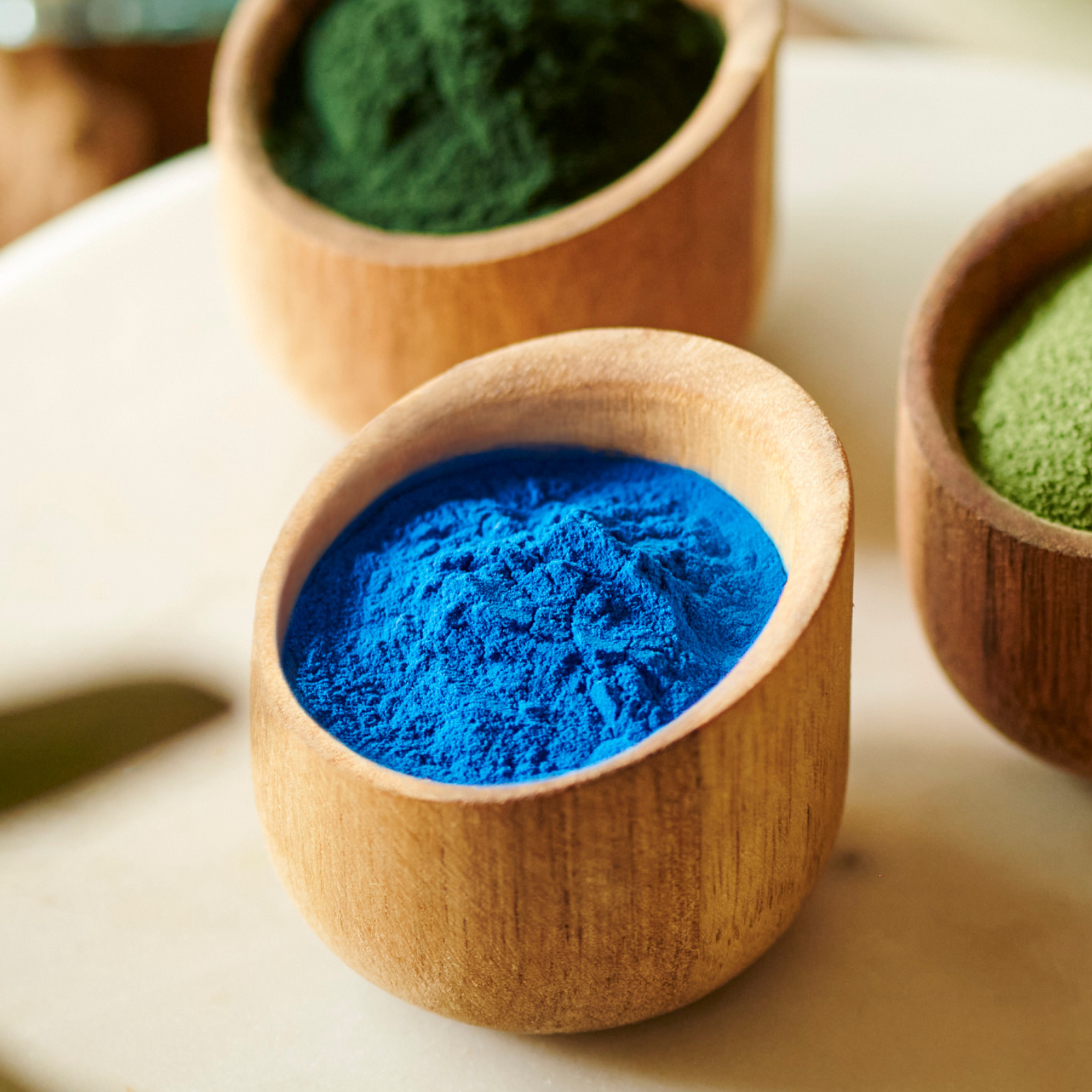 Three small wooden bowls containing colored powders; one with blue, and two with green powder, arranged on a light surface.