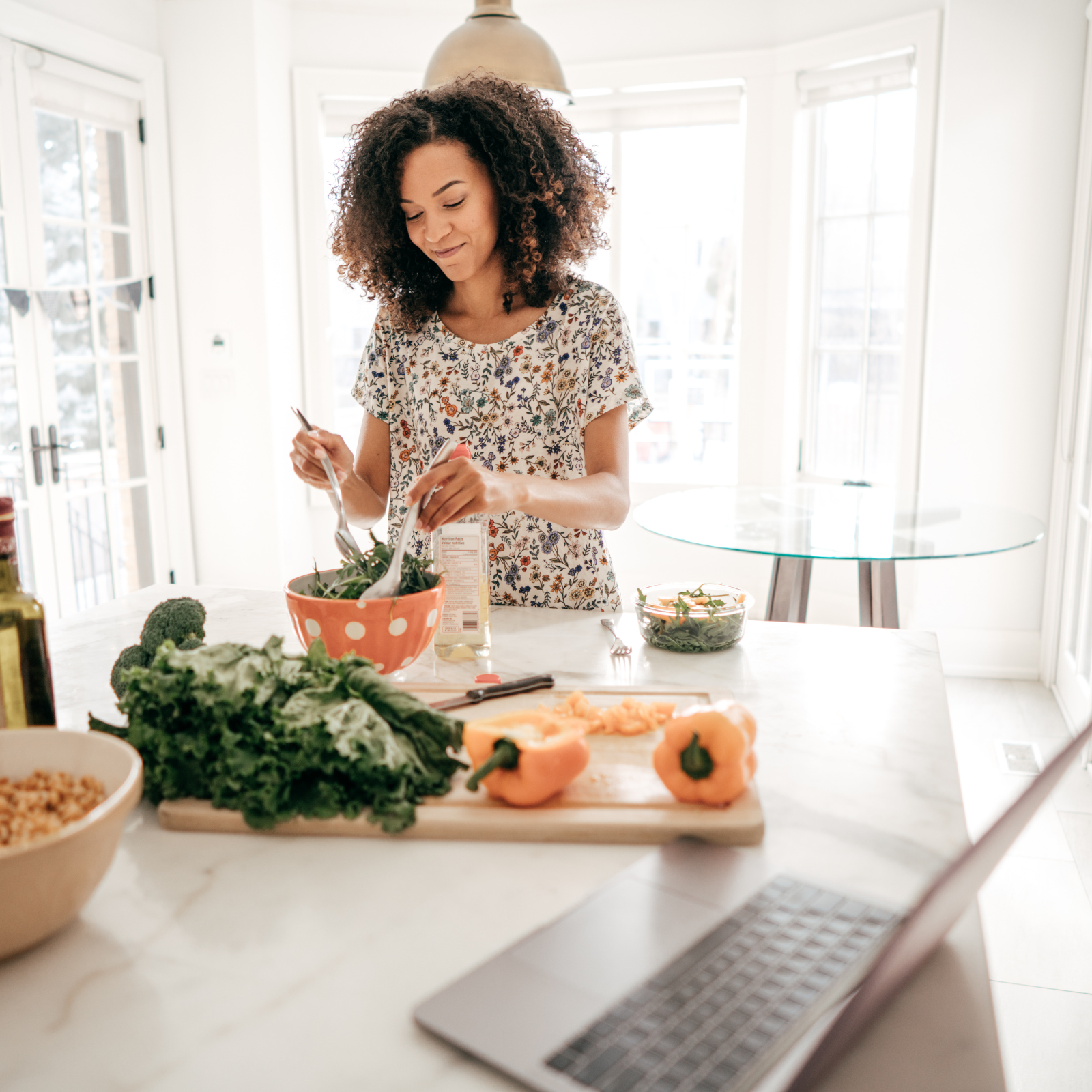 A woman with curly hair preparing a salad in a bright kitchen with large windows. There are vegetables such as yellow bell peppers, broccoli, and mixed greens on the counter, along with a laptop and salad bowls.