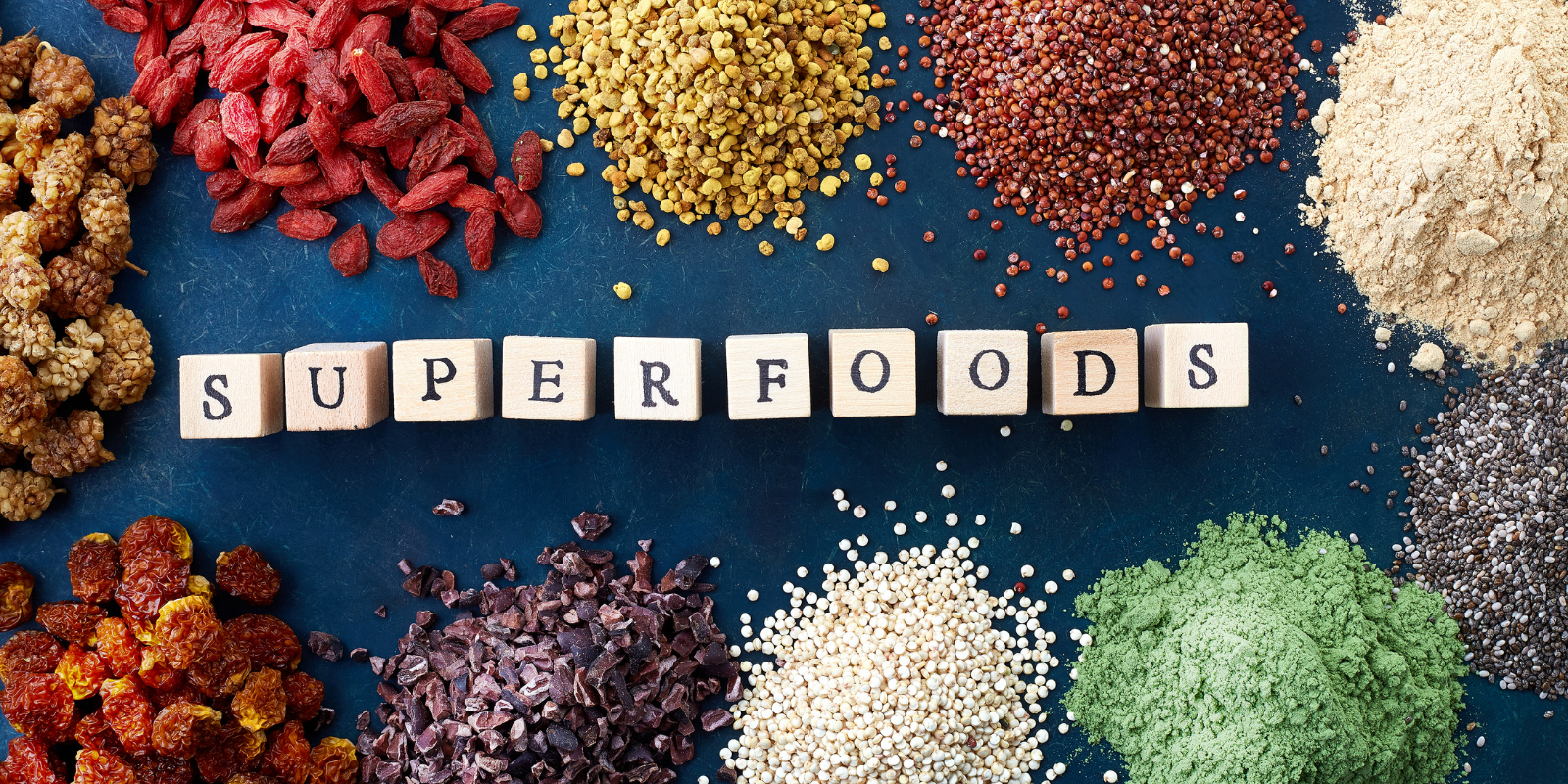 Various superfoods such as dried goji berries, mulberries, bee pollen, quinoa, ground powders, chia seeds, and other colorful healthy ingredients arranged on a blue surface with the words 'SUPERFOODS' spelled out in wooden blocks.