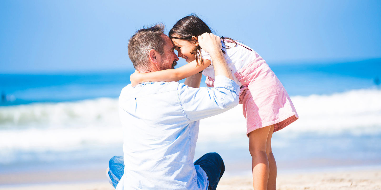 A man and young girl at the beach with their foreheads touching, smiling and holding each other's faces, with ocean waves in the background.