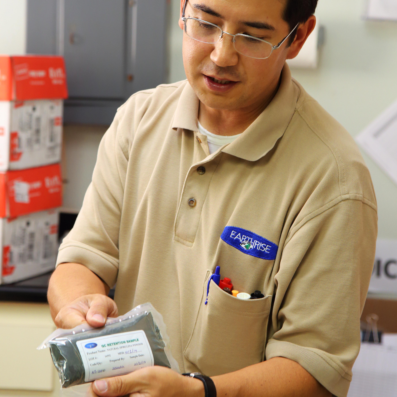 A man in a beige polo shirt with an 'Earthrise' logo on the chest, holding a sealed package labeled 'QC Retention Sample'.