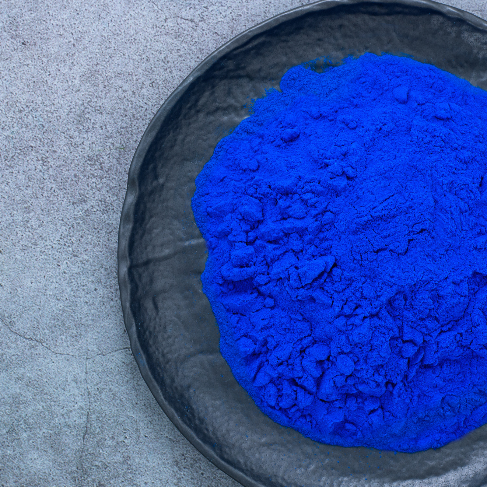 Bright blue powdered pigment in a black bowl on a gray textured surface.