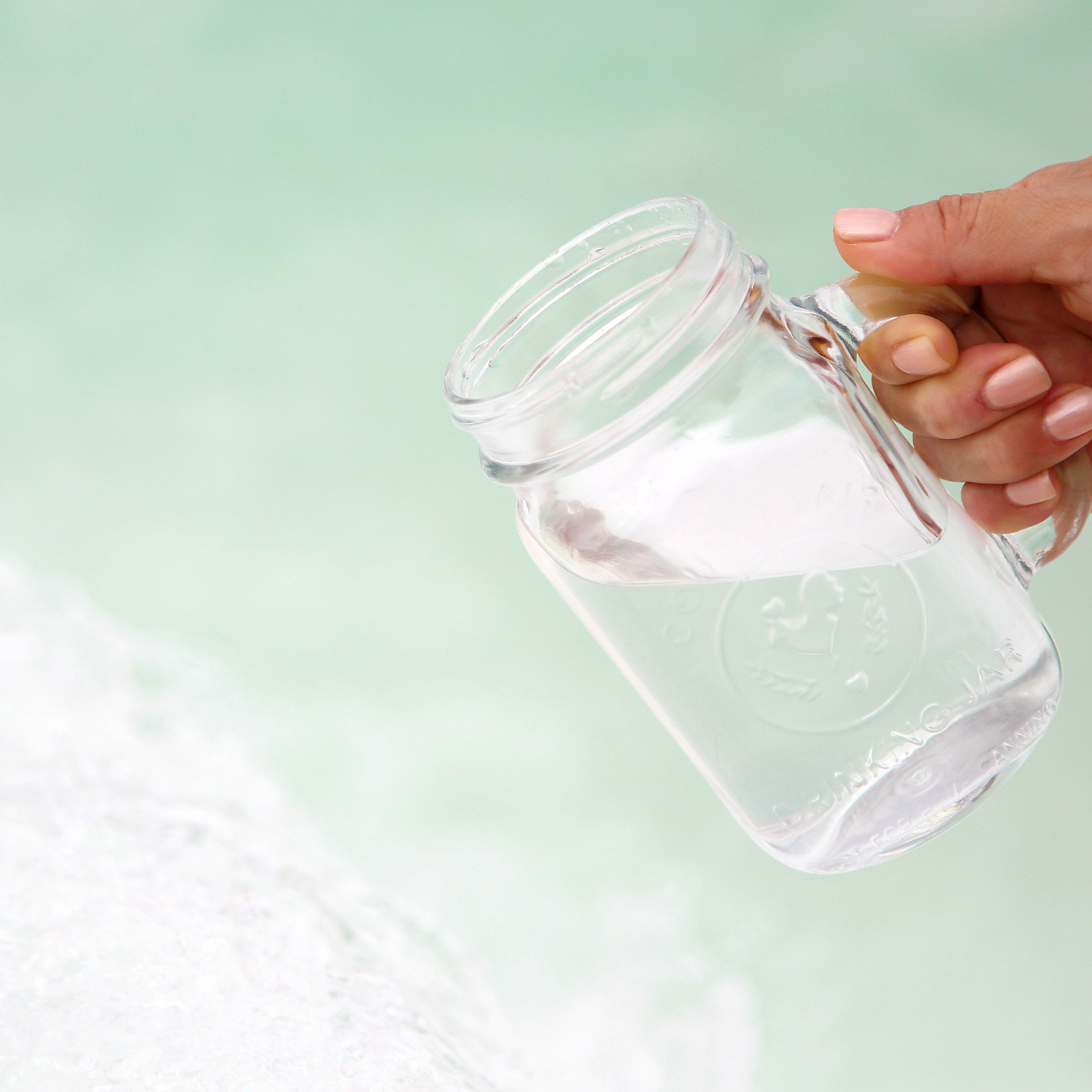 Hand holding a glass Mason jar tilted to pour water into a light green background