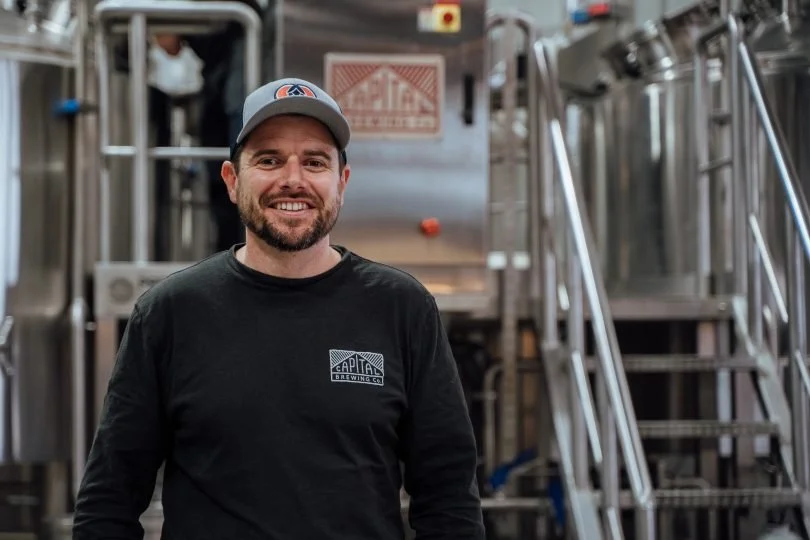 A man smiling in a brewery, wearing a baseball cap and a black long-sleeve shirt with a brewery logo, standing in front of brewing equipment.