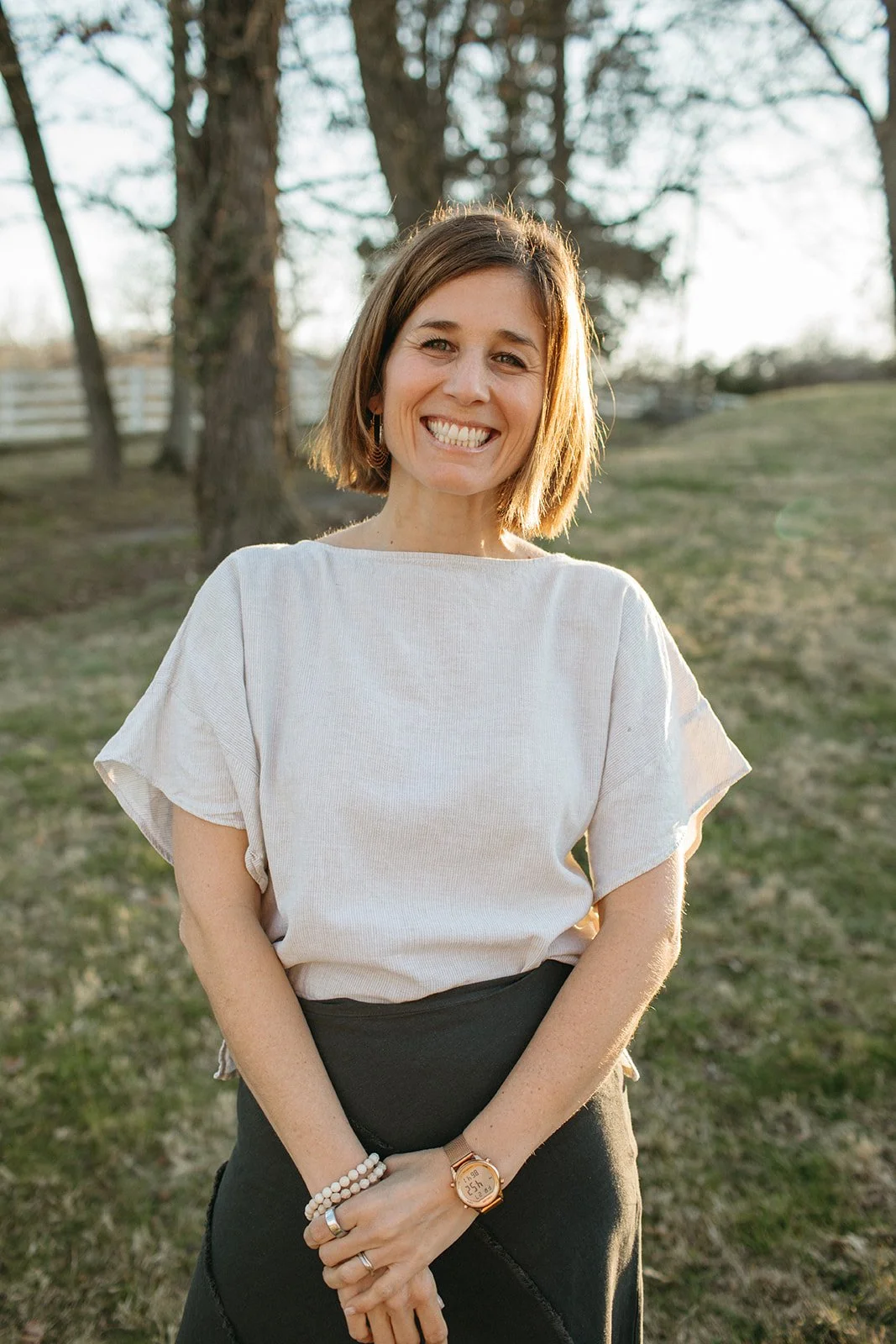 A woman standing outdoors in a grassy area with trees, smiling at the camera during sunset, wearing a beige top, black skirt, beaded bracelet, and a watch.