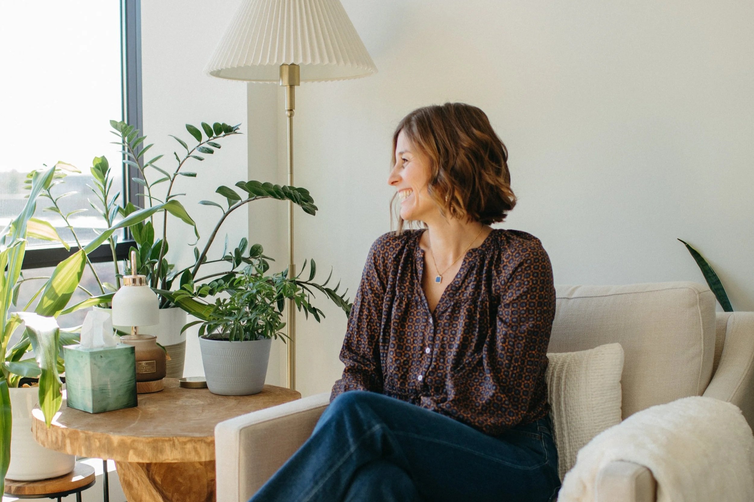 A woman with short wavy hair sitting on a beige sofa, smiling and looking out the window. She is wearing a patterned blouse and a necklace. There are green potted plants and a lamp on a wooden side table next to her.