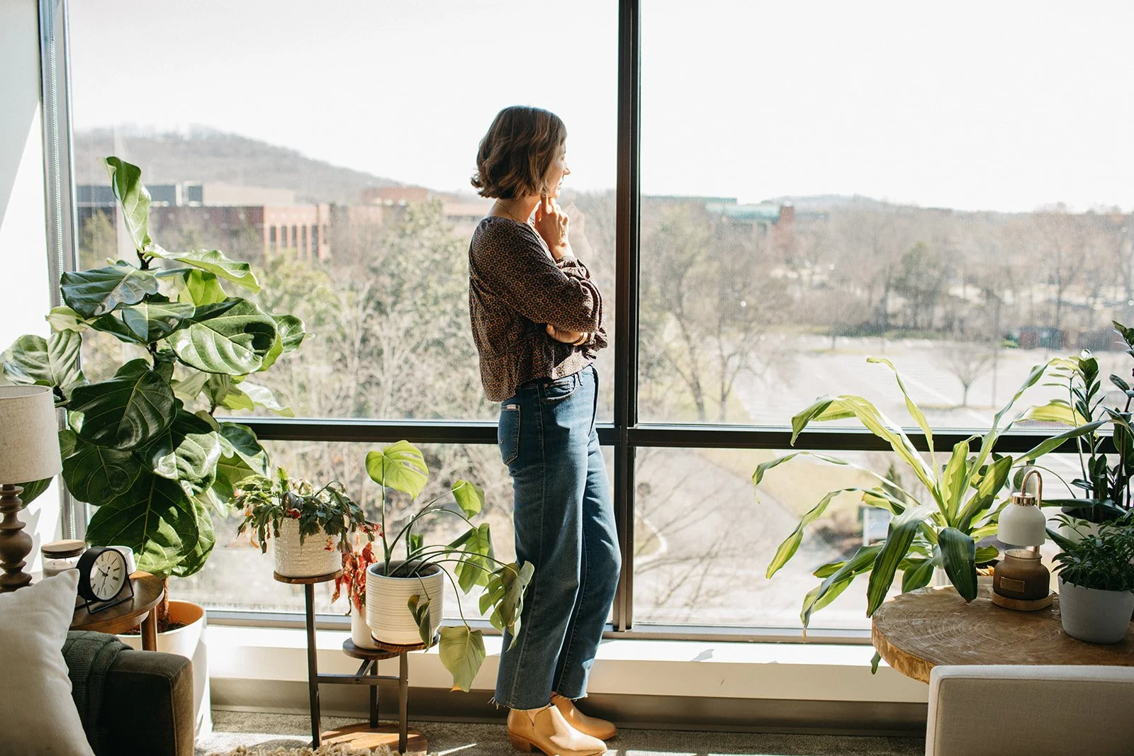 Woman standing by a large window with indoor plants on side tables and a couch nearby, looking outside.