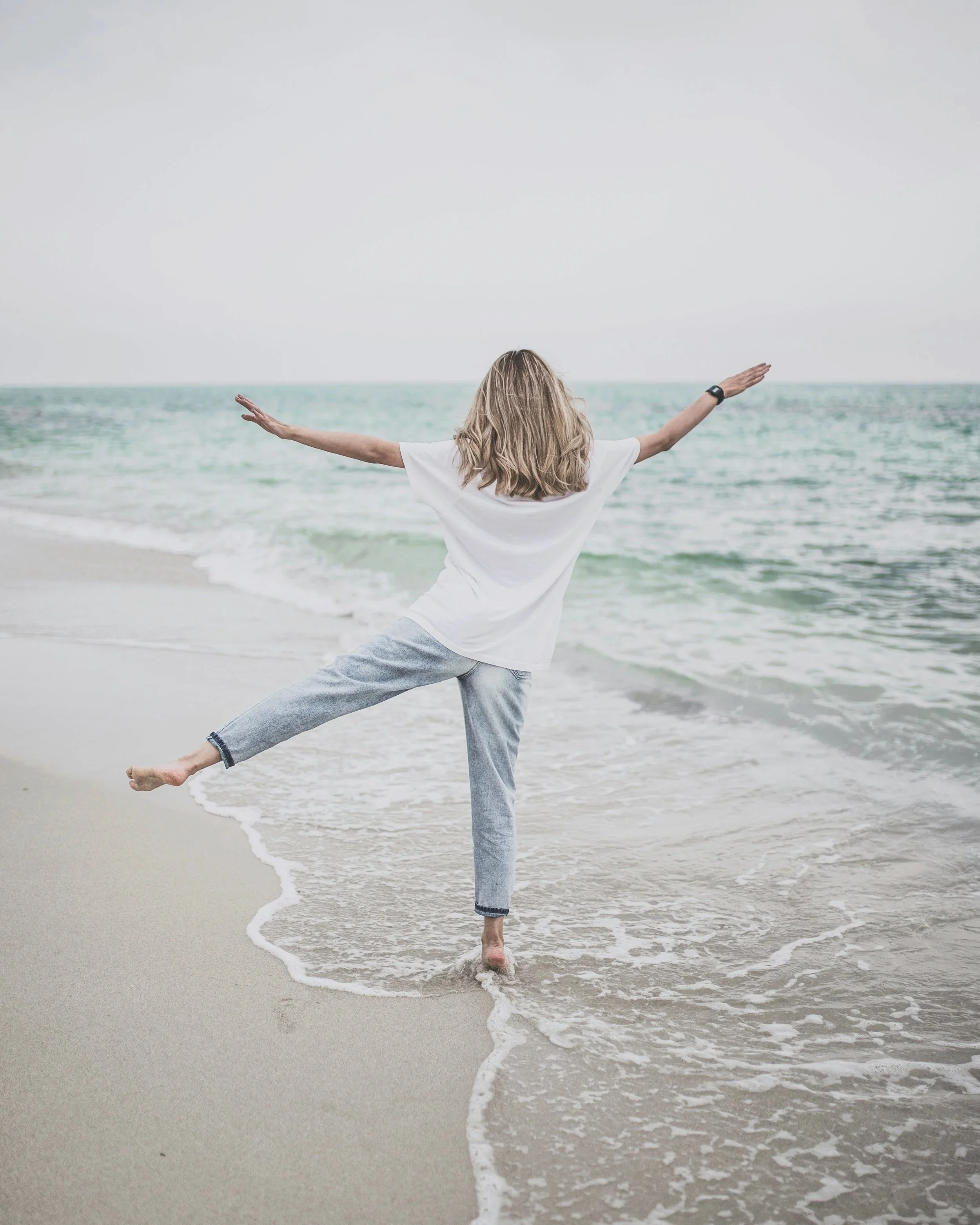 A woman with blonde hair, wearing a white t-shirt and gray sweatpants, standing on a beach with arms outstretched and one foot lifted, facing the ocean under an overcast sky.