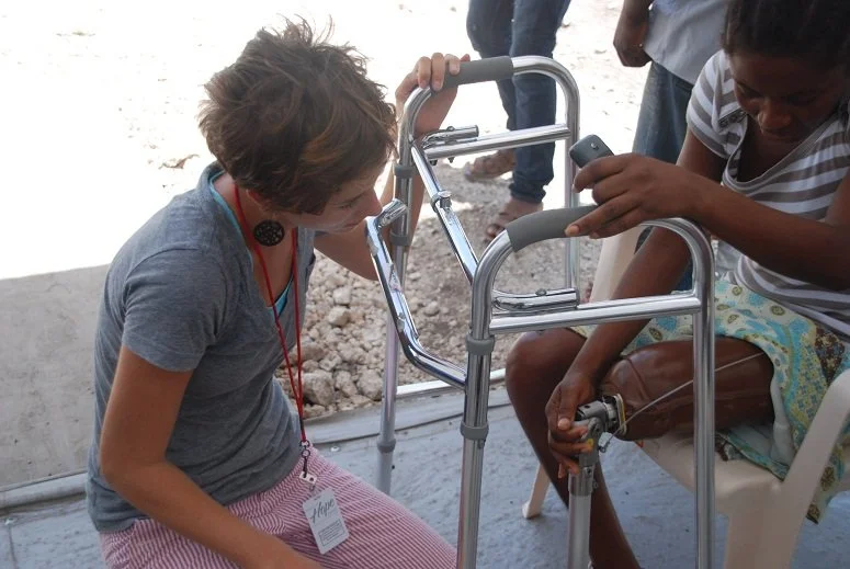 A woman with short hair wearing a gray shirt and pink striped pants assists a woman with dark hair, wearing a striped shirt and colorful skirt, at a rural or outdoor setting with a walker.