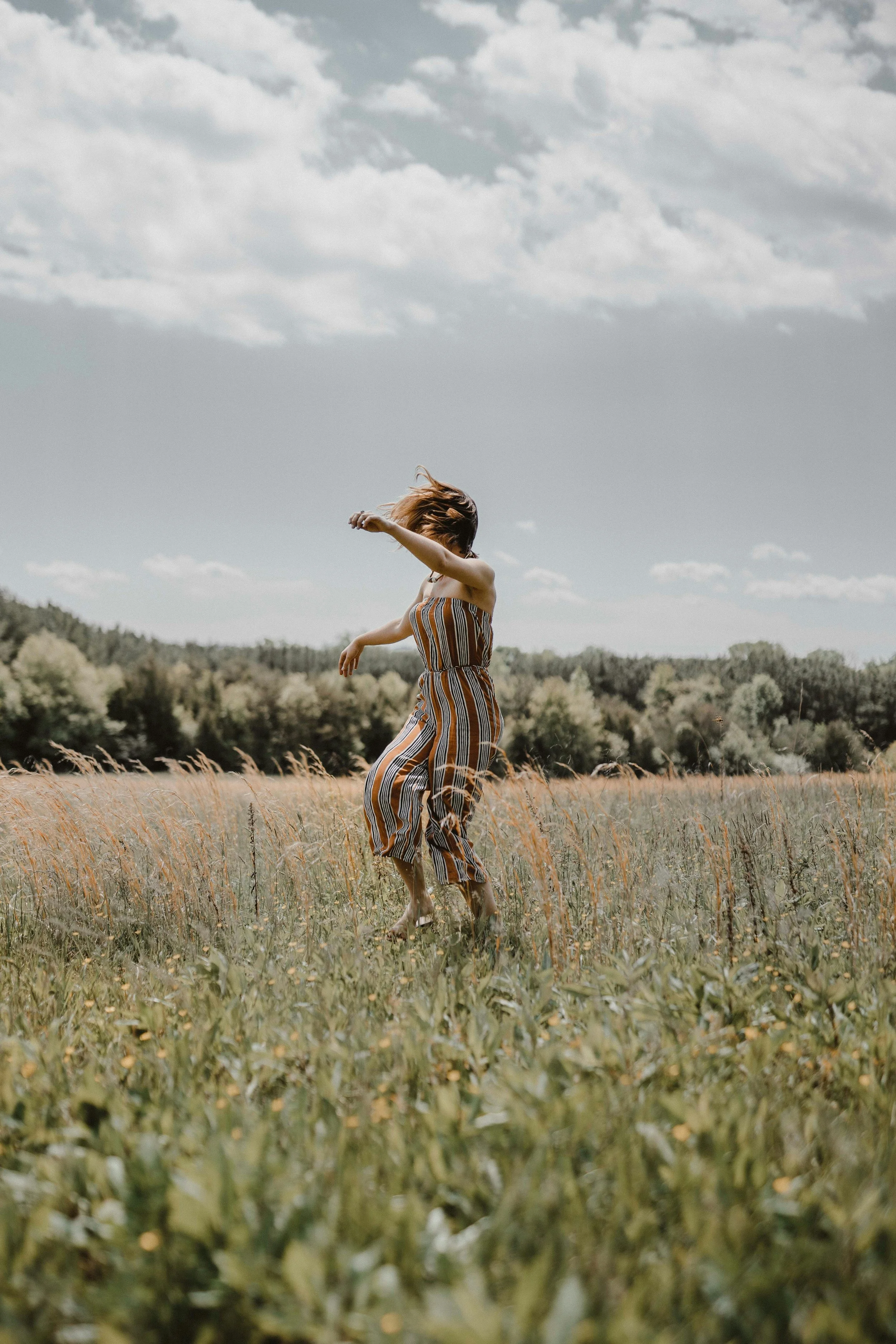A person with long hair wearing a striped jumpsuit is dancing in a field of tall grass with trees and hills in the background under a cloudy sky.