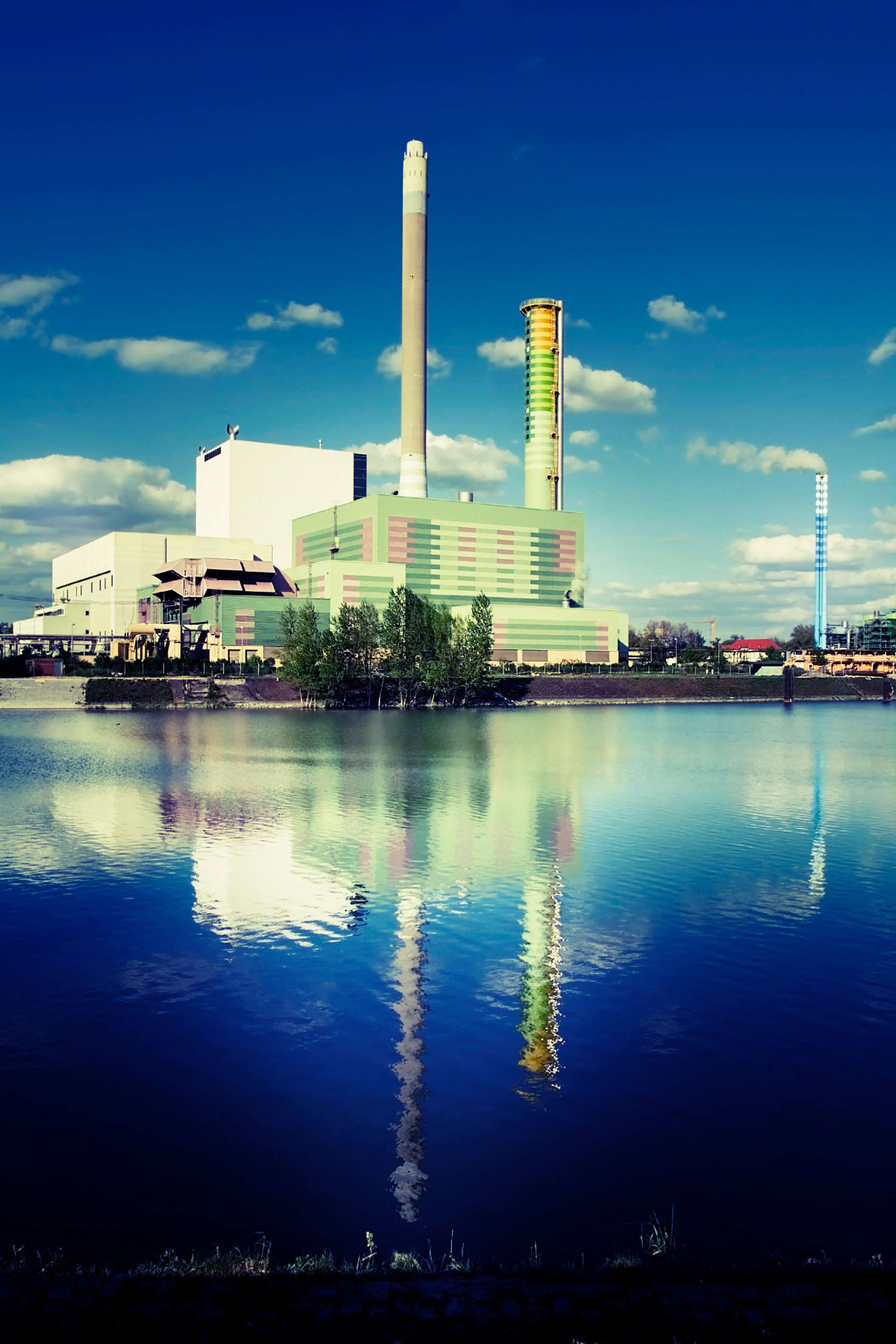 View of a large power plant with tall smokestacks reflected in a river under a partly cloudy sky.