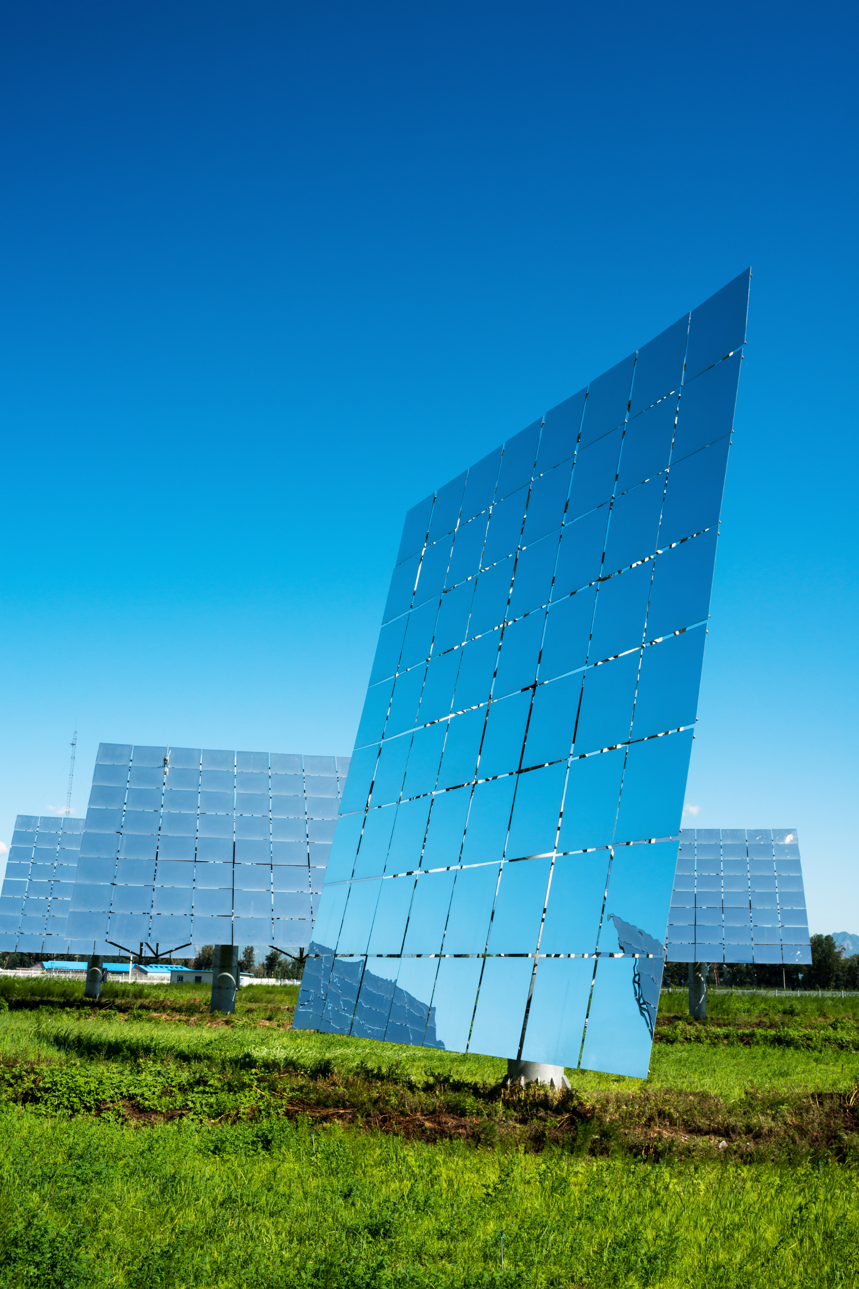 Multiple large solar panels in a field, reflecting the sky and clouds in bright, clear weather.