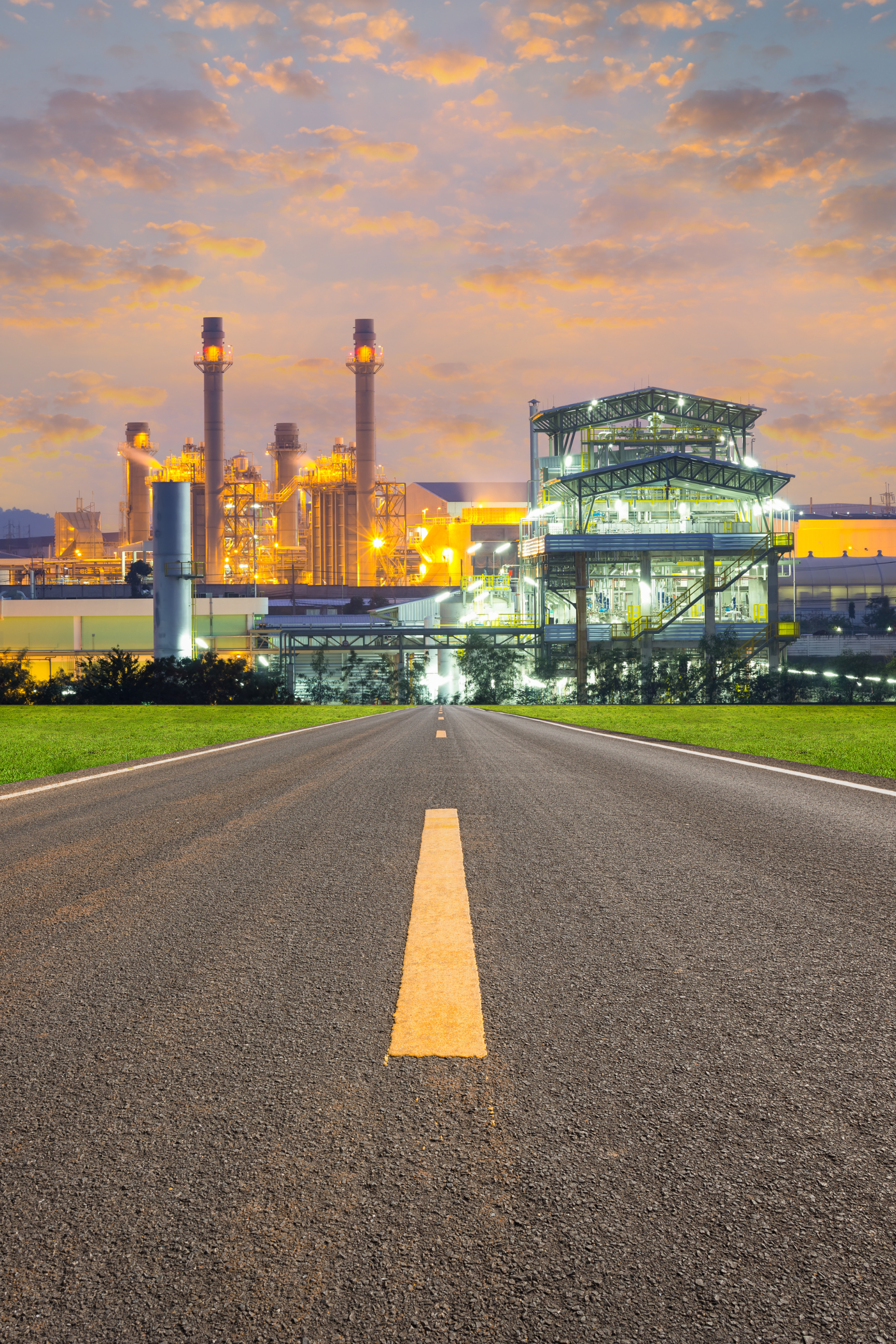A straight road leading to an industrial factory at sunset, with orange sky and clouds in the background.