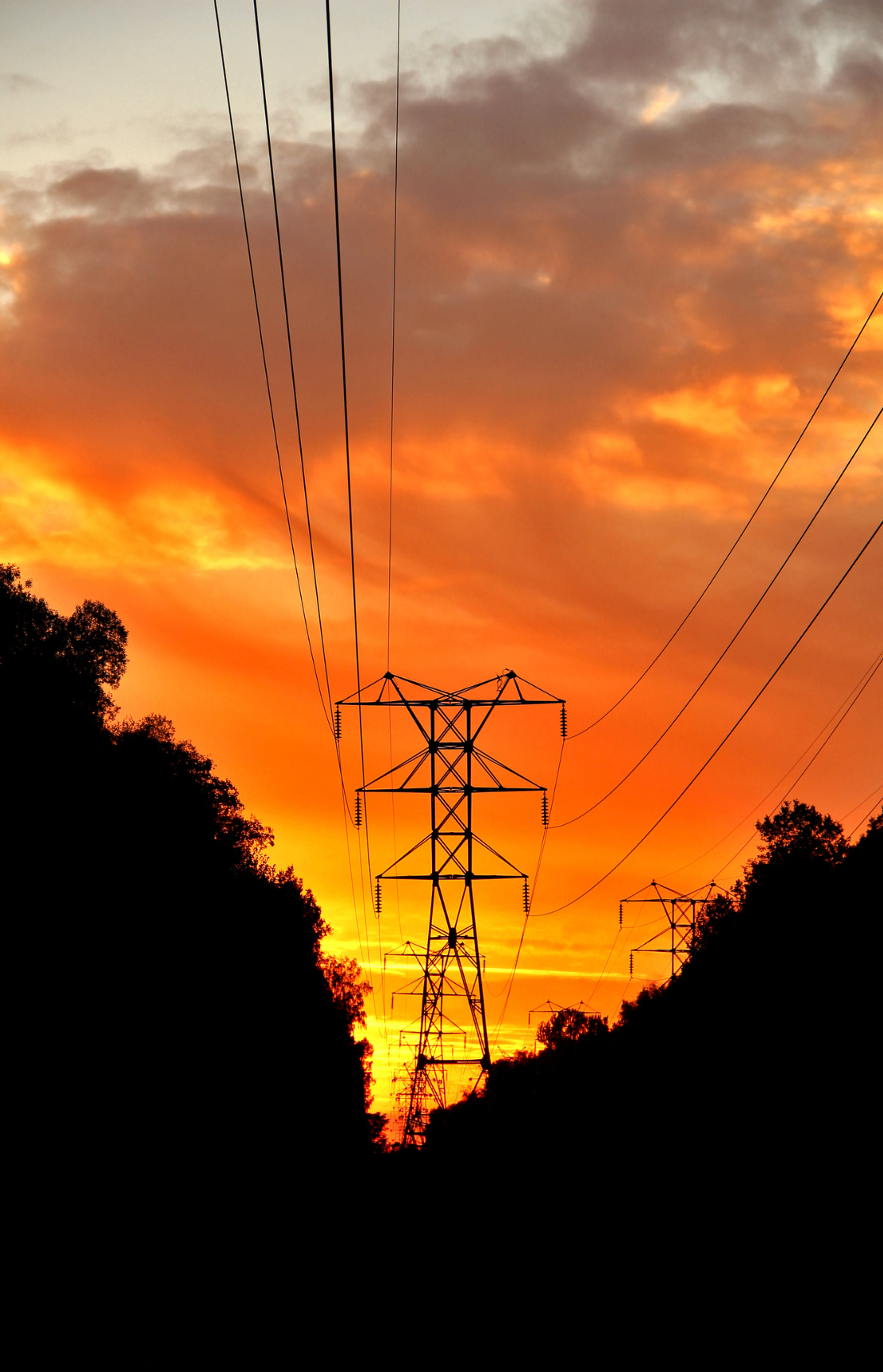 Silhouettes of power line towers against a vibrant orange sunset with clouds.