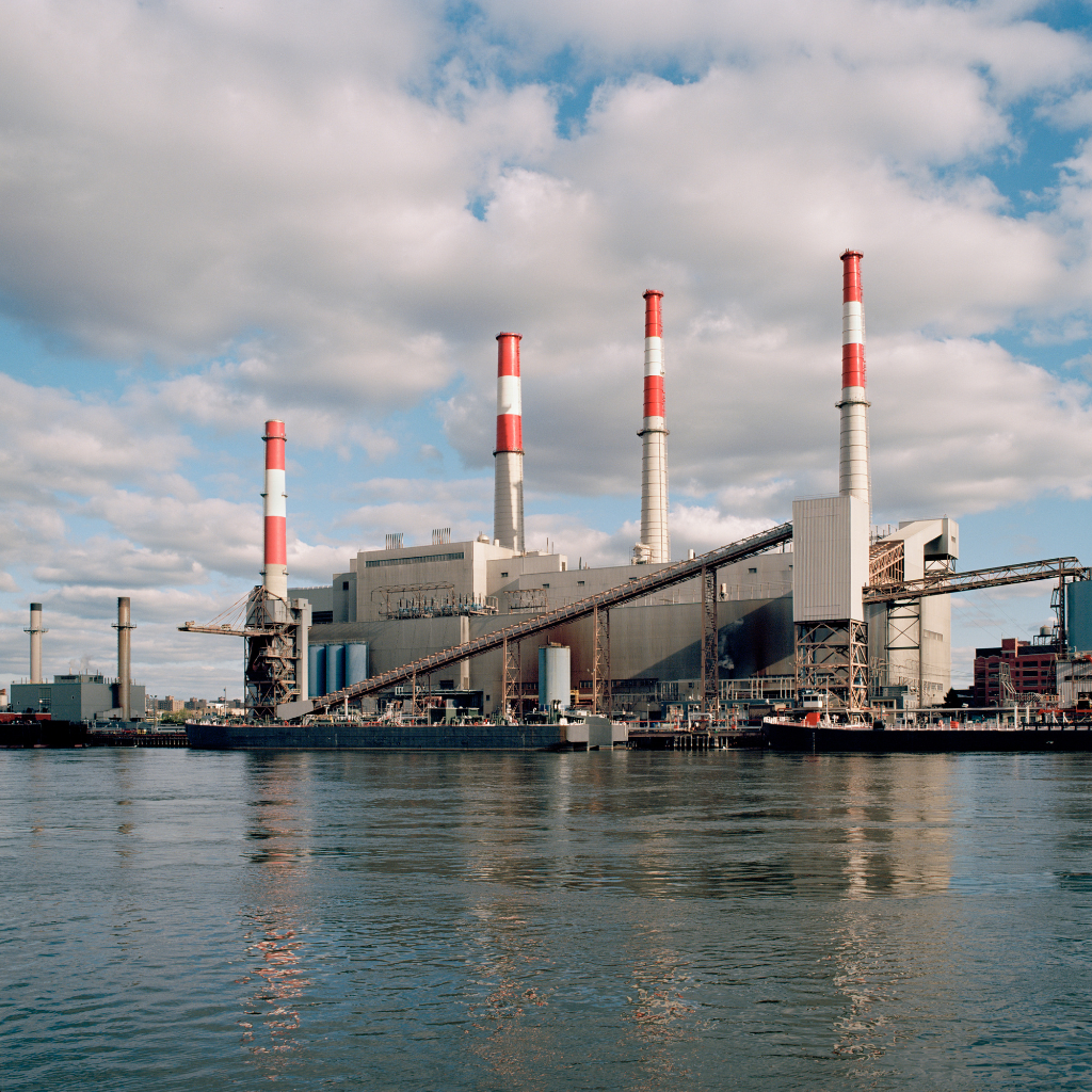 A large industrial power plant with four tall smoke stacks painted with red and white stripes, situated along a waterfront with calm water reflecting the structure, partly cloudy sky overhead.