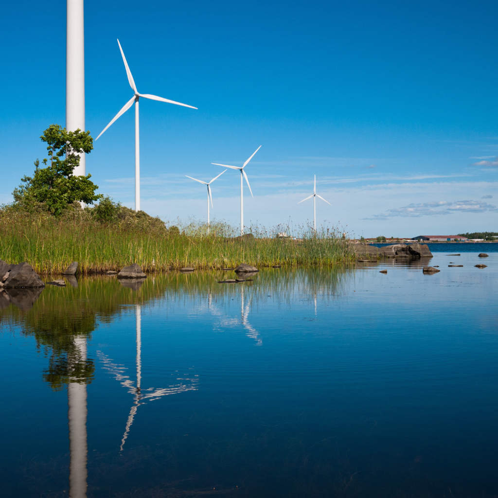 Multiple white wind turbines along a green shoreline near a calm body of water, with a clear blue sky above.
