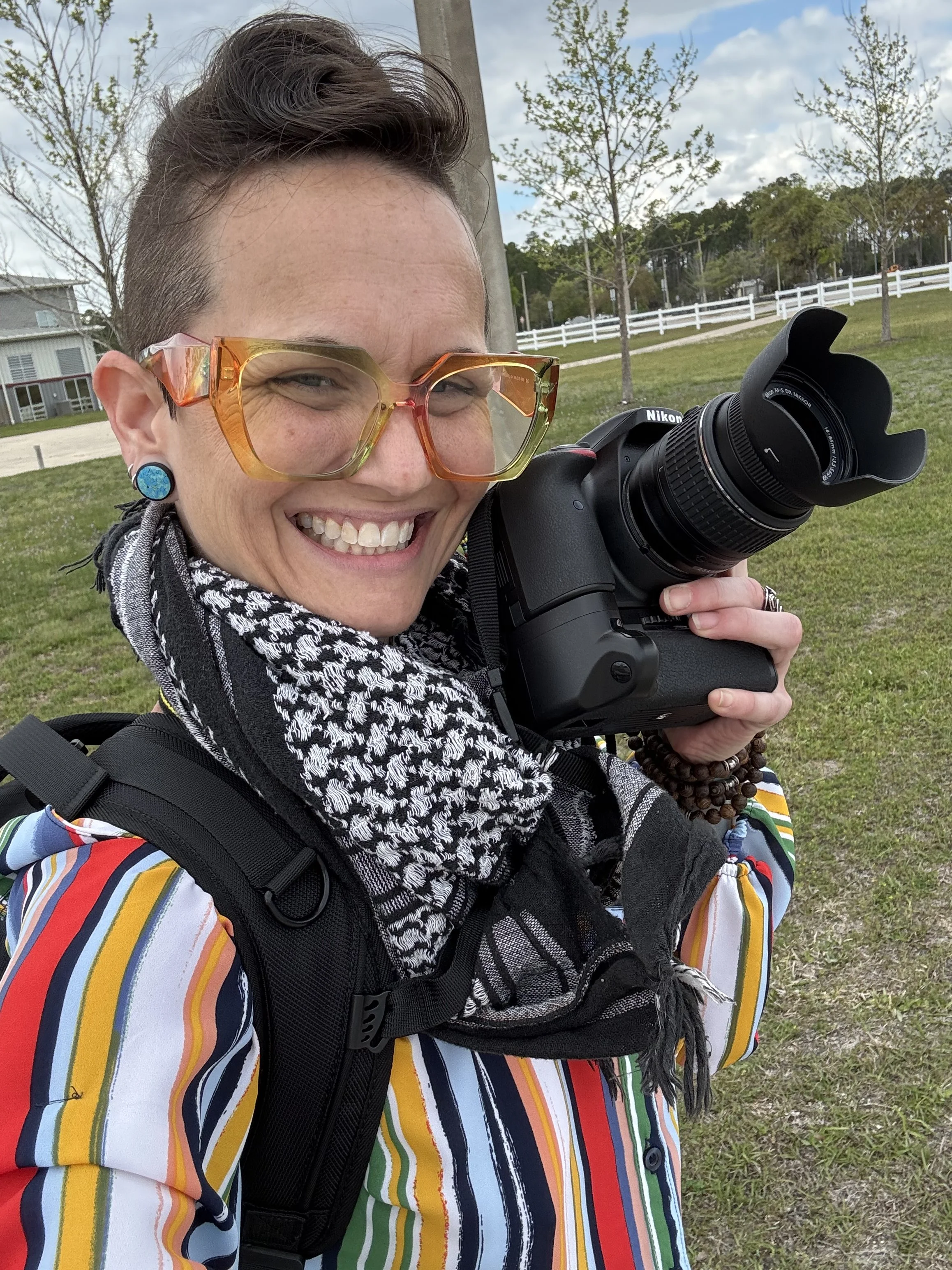 A woman with short hair, colorful glasses, and blue earrings is smiling and holding a Nikon camera with a large lens. She is wearing a multicolored striped jacket, a black and white patterned scarf, and a backpack, standing outdoors in a park with trees and a white fence in the background.