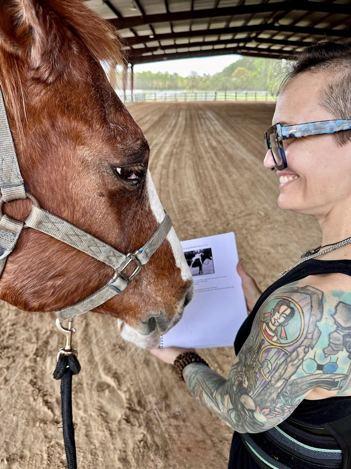 A woman with tattoos and glasses smiling at a brown and white horse inside a covered riding arena.