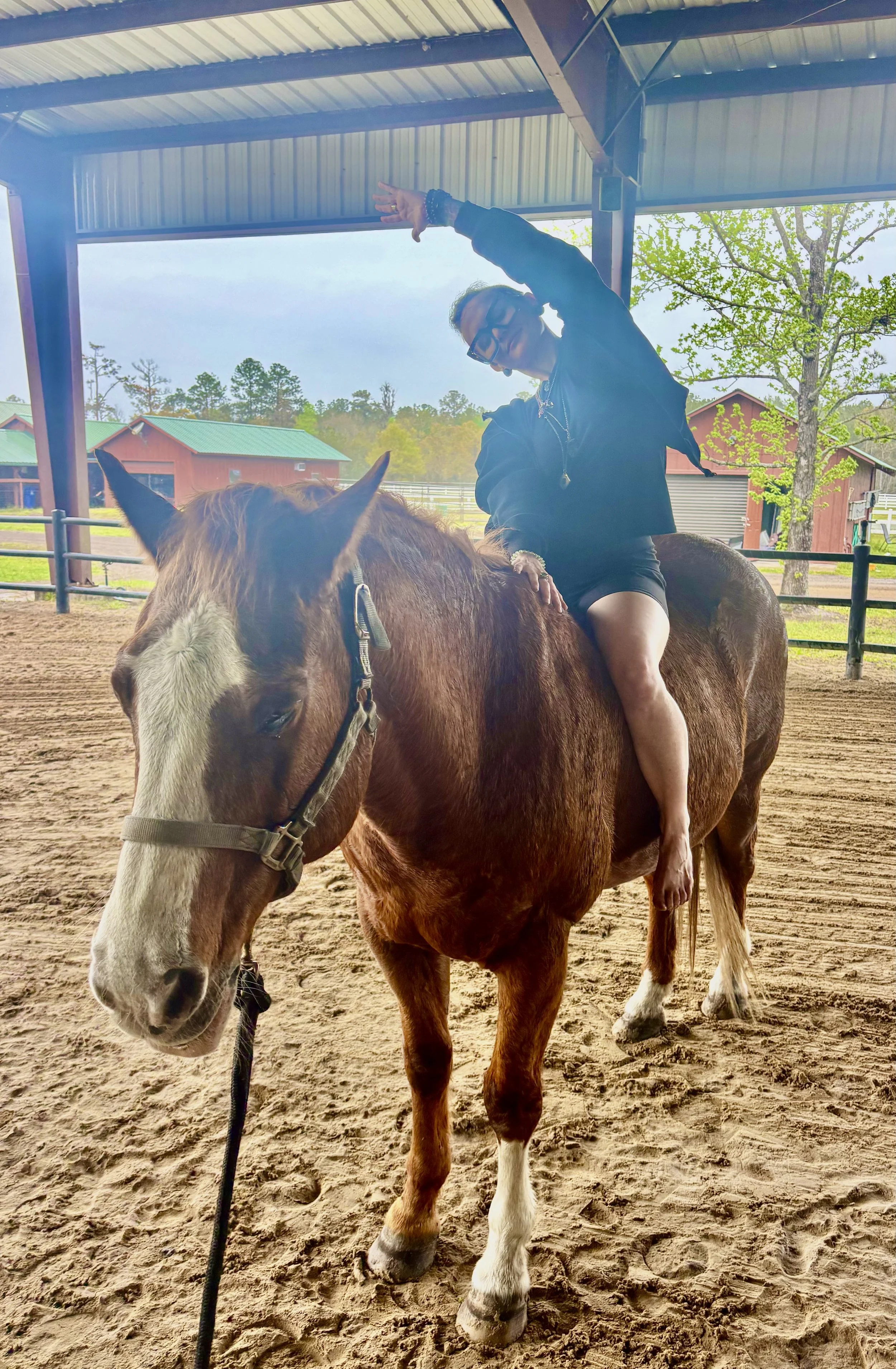 A woman riding a brown and white horse inside a covered riding arena, with barns and trees in the background.