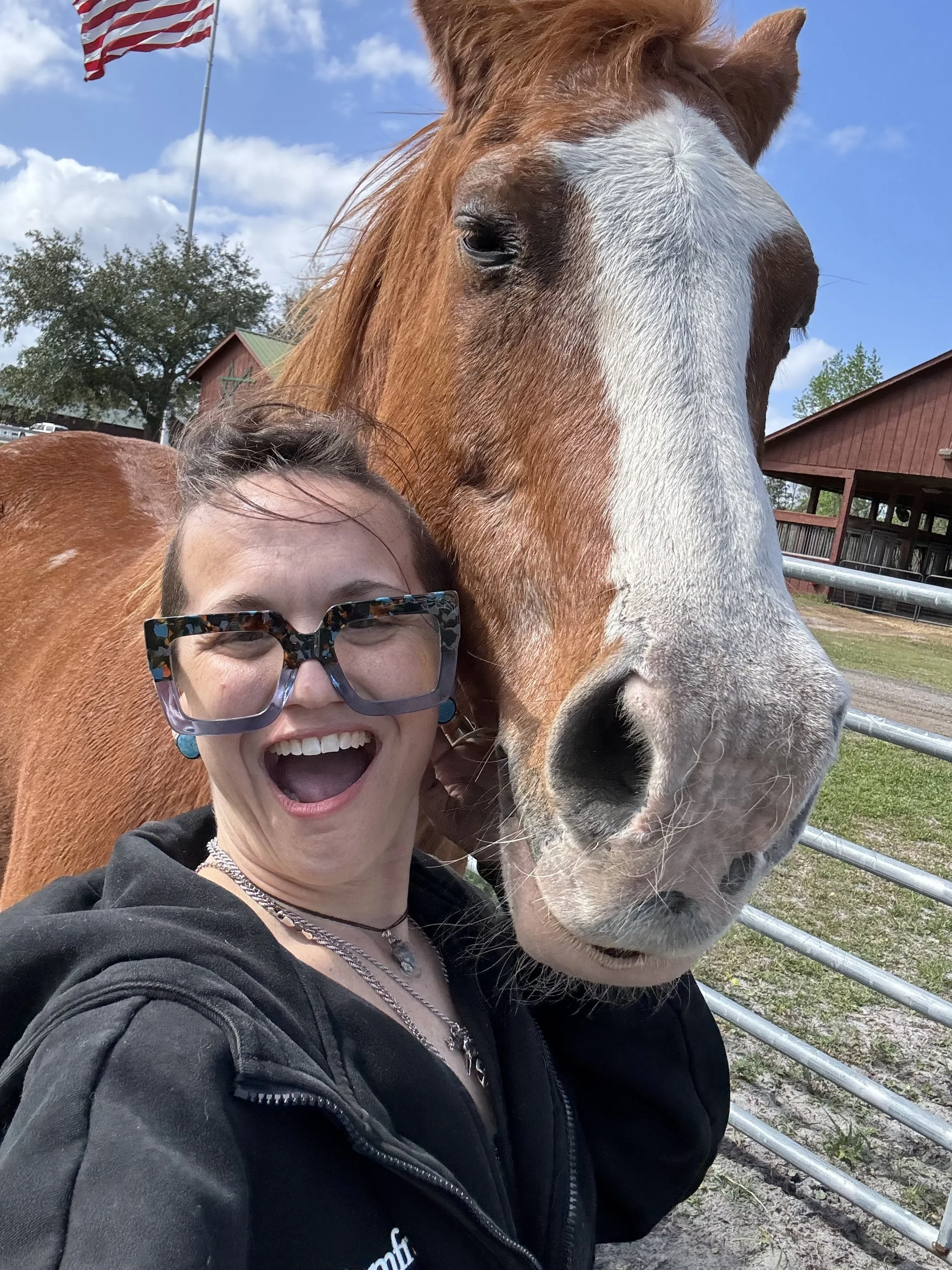 A woman smiling and joyfully taking a selfie with a large brown and white horse at a farm or equestrian facility.