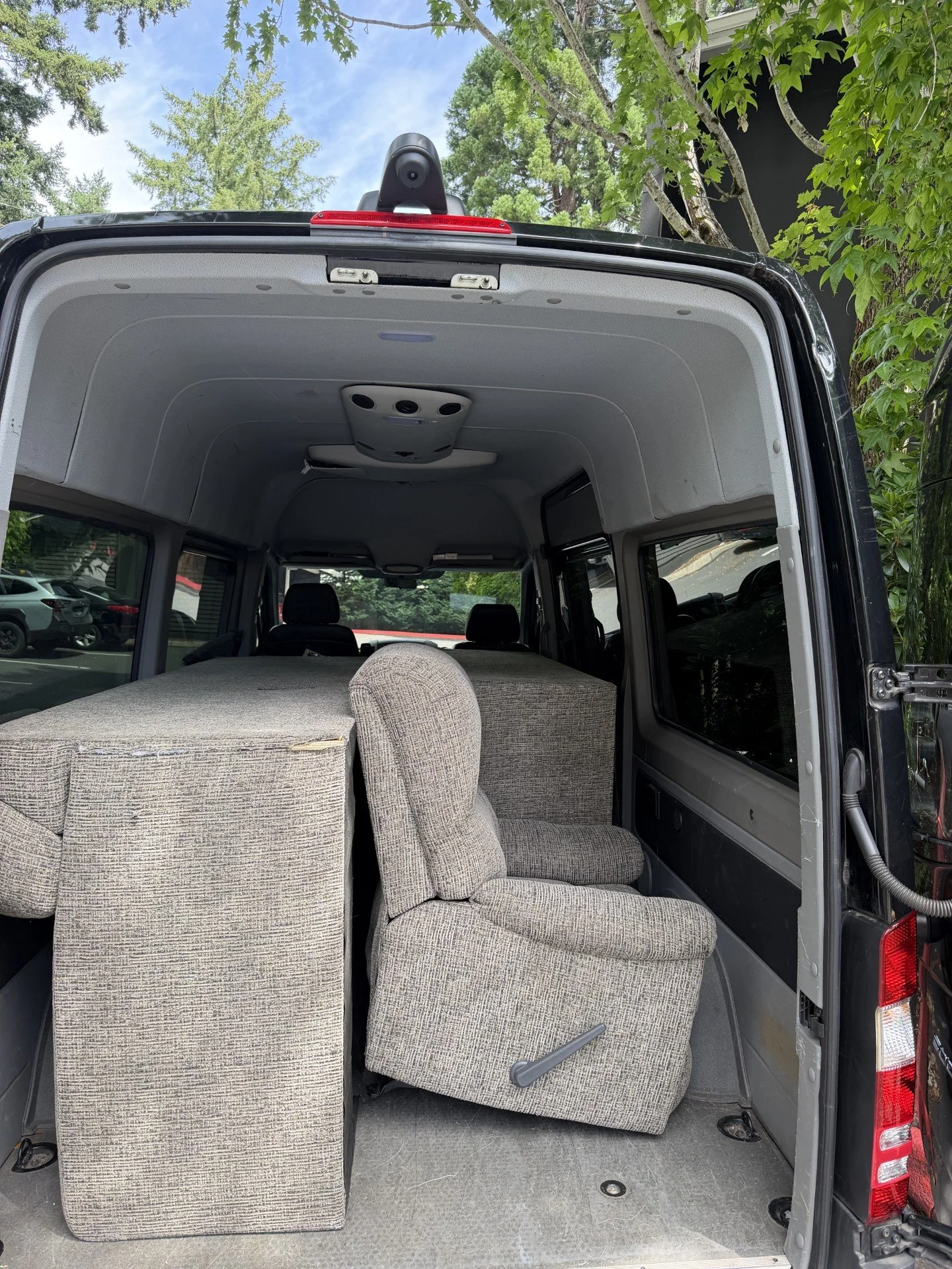 Interior of a vehicle with gray upholstered seats and a flat, carpeted surface, viewed from the back with the rear door open, set outdoors with trees and parked cars visible through the windows.