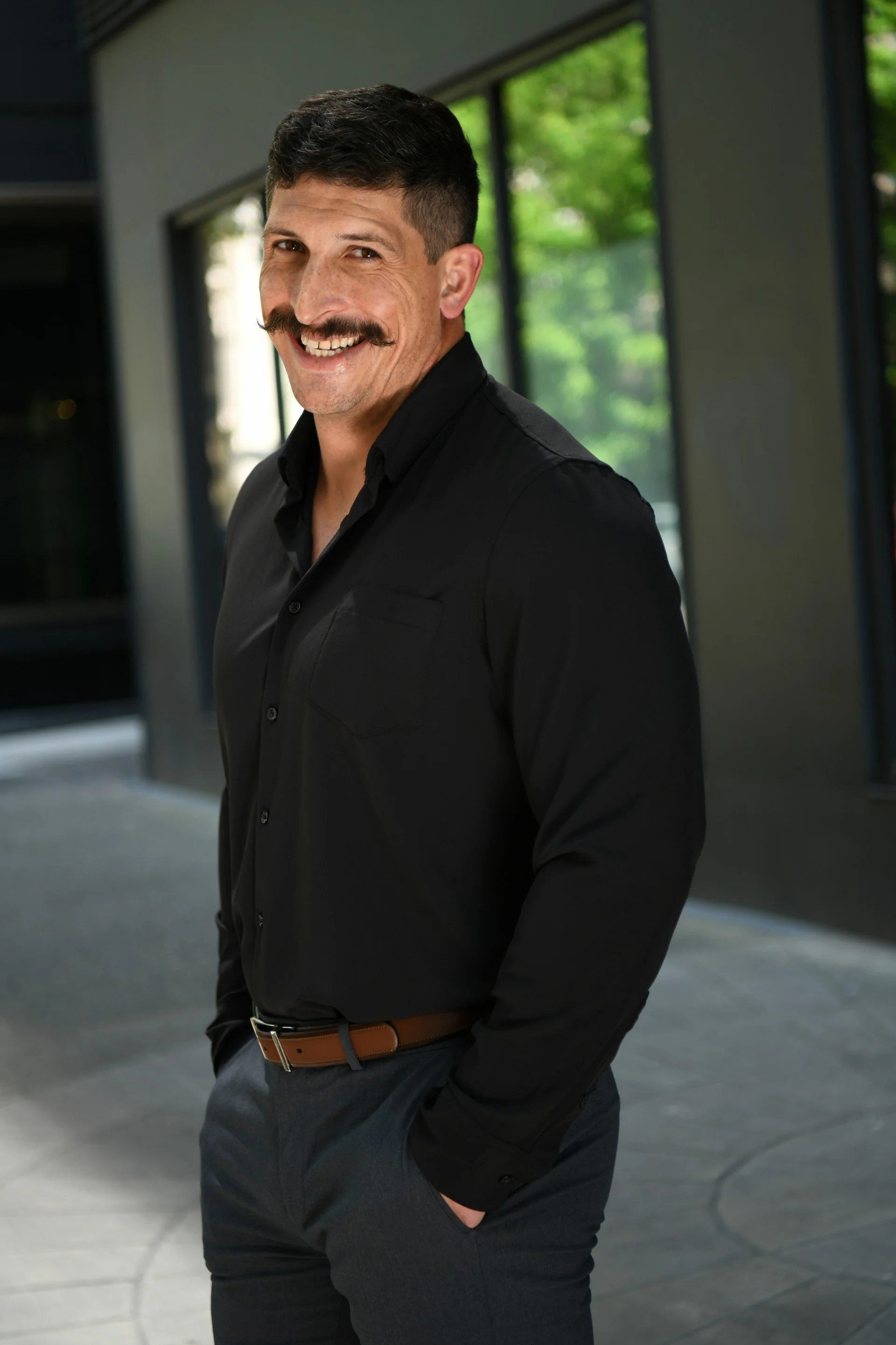 A smiling man with a mustache and short dark hair poses in a modern office building, wearing a black shirt and gray pants with his hands in his pockets.