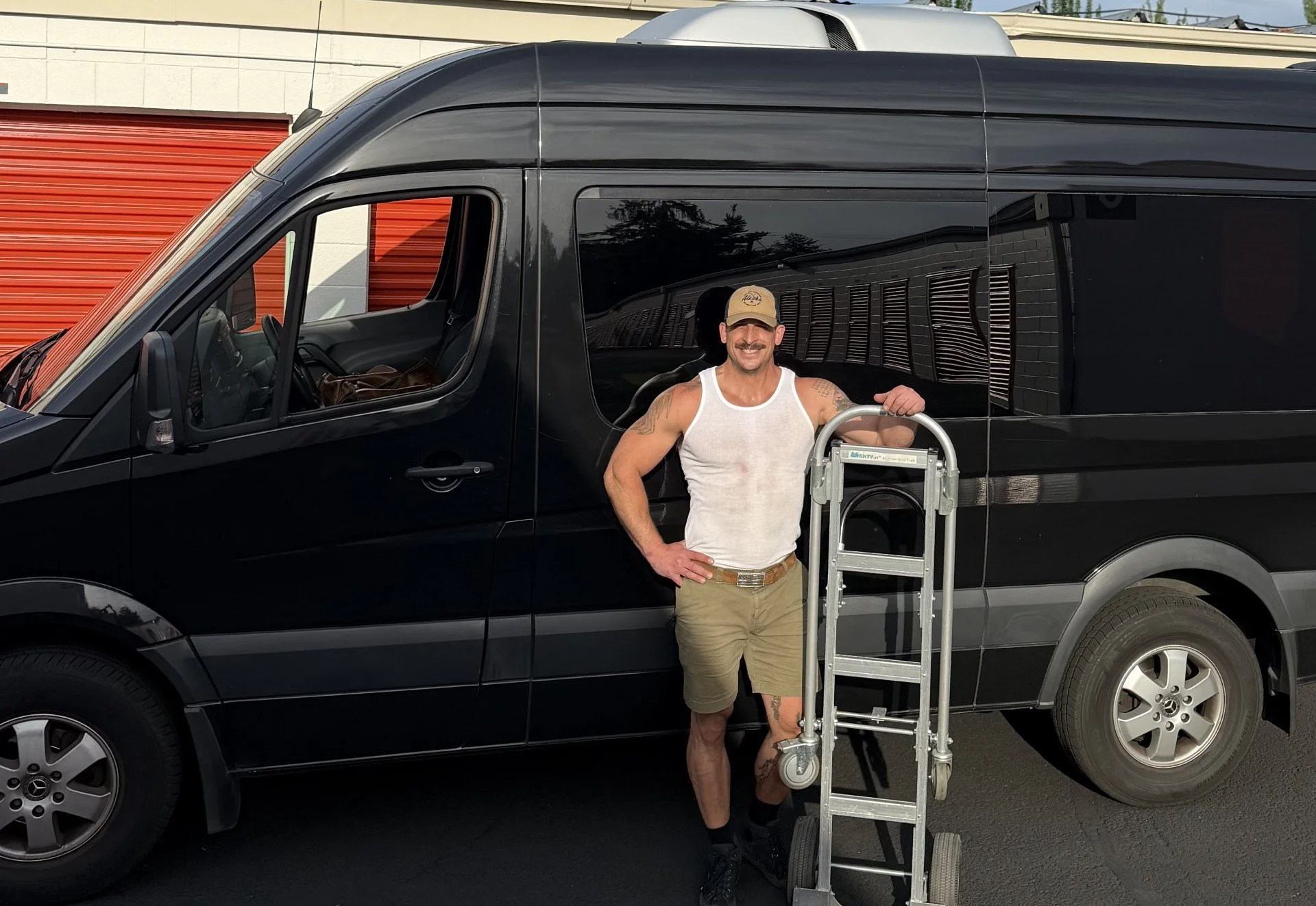 A man wearing a white tank top, tan shorts, and a beige cap standing next to a large black van with a ladder and a grabber device, smiling at the camera in a parking lot.