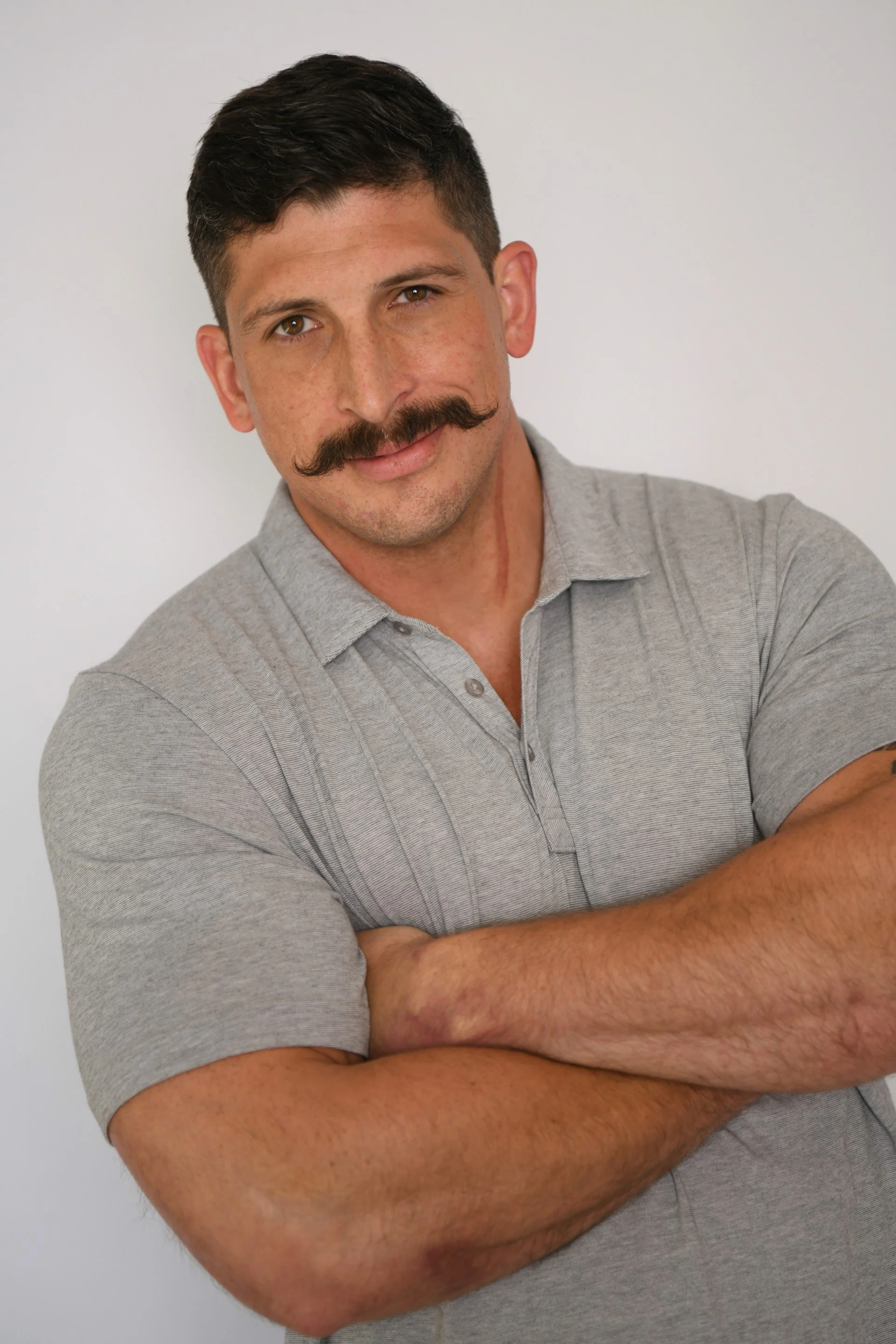 A man with a dark mustache, short dark hair, and light skin, wearing a light gray polo shirt with his arms crossed, standing against a plain white background.