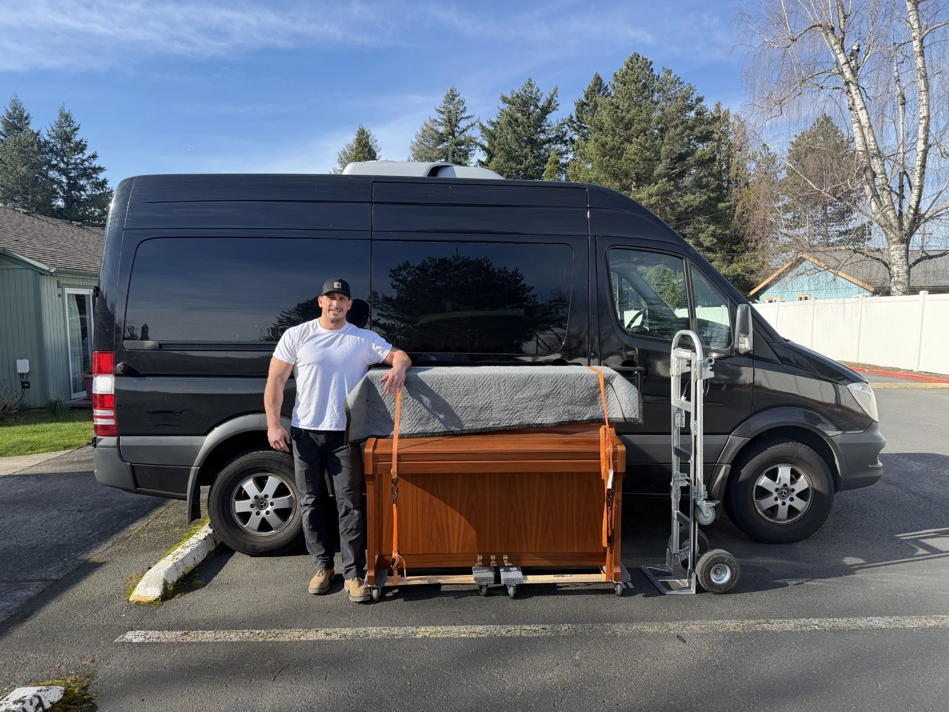 A man standing beside a black van with moving supplies, including furniture and a dolly, parked in a residential area on a sunny day.