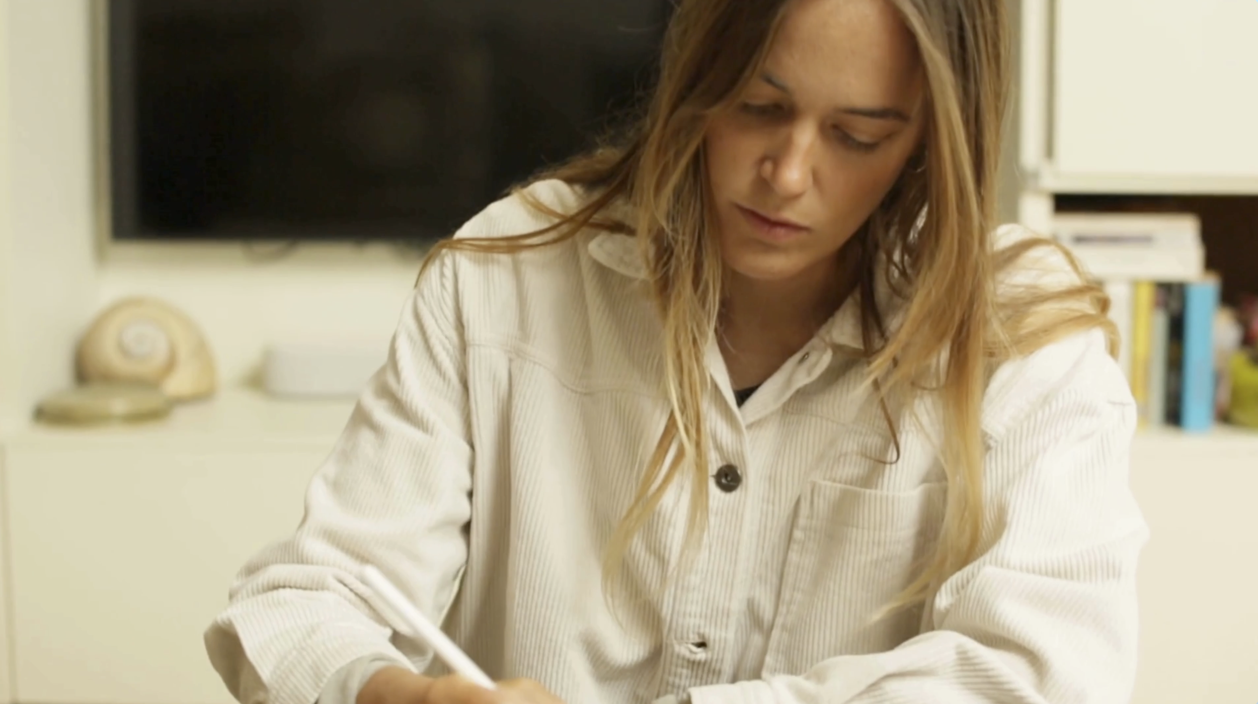 Una donna con capelli castano chiaro che indossa una camicia bianca con righe e scrive, seduta a una scrivania.