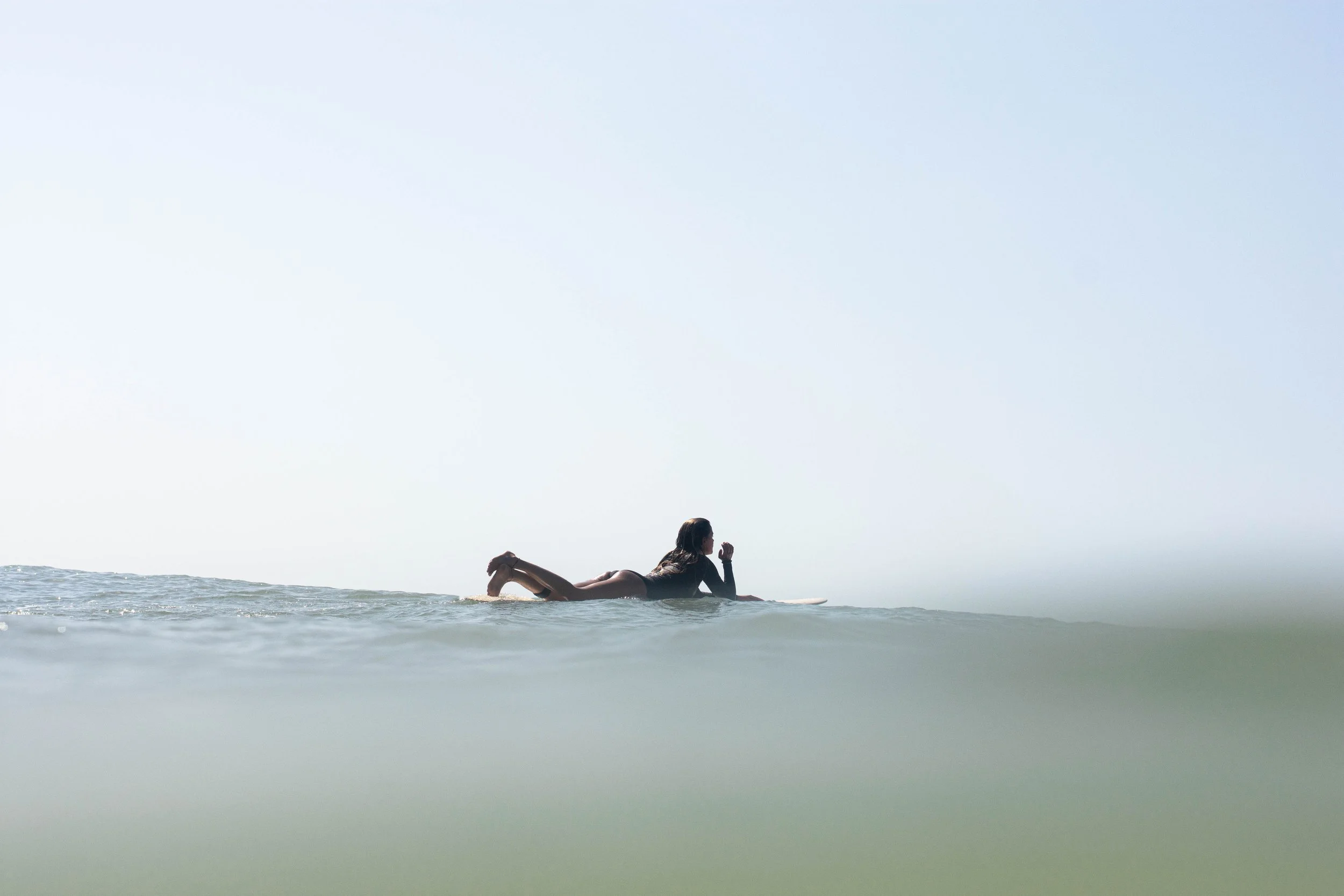 Una donna distesa sull'acqua mentre fa surf, con vista del mare e il cielo sereno.