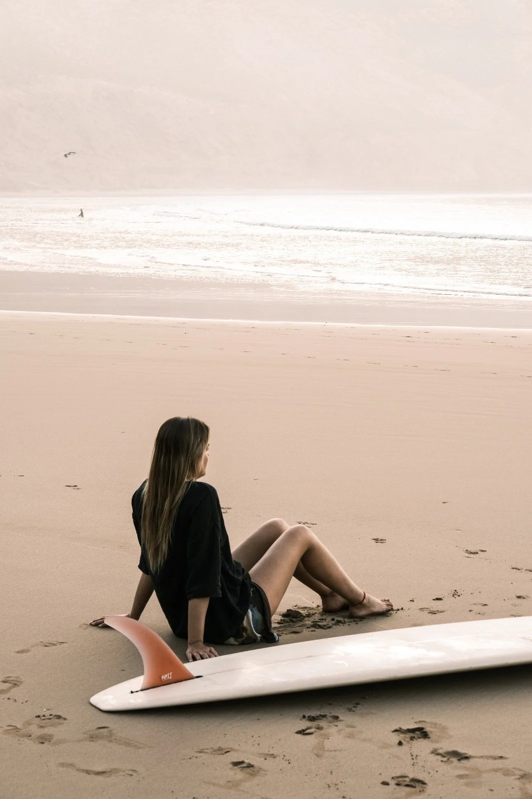 Una ragazza seduta sulla spiaggia vicino a una tavola da surf con la pinna, guarda l'oceano in lontananza. La scena è serena e il cielo è coperto.