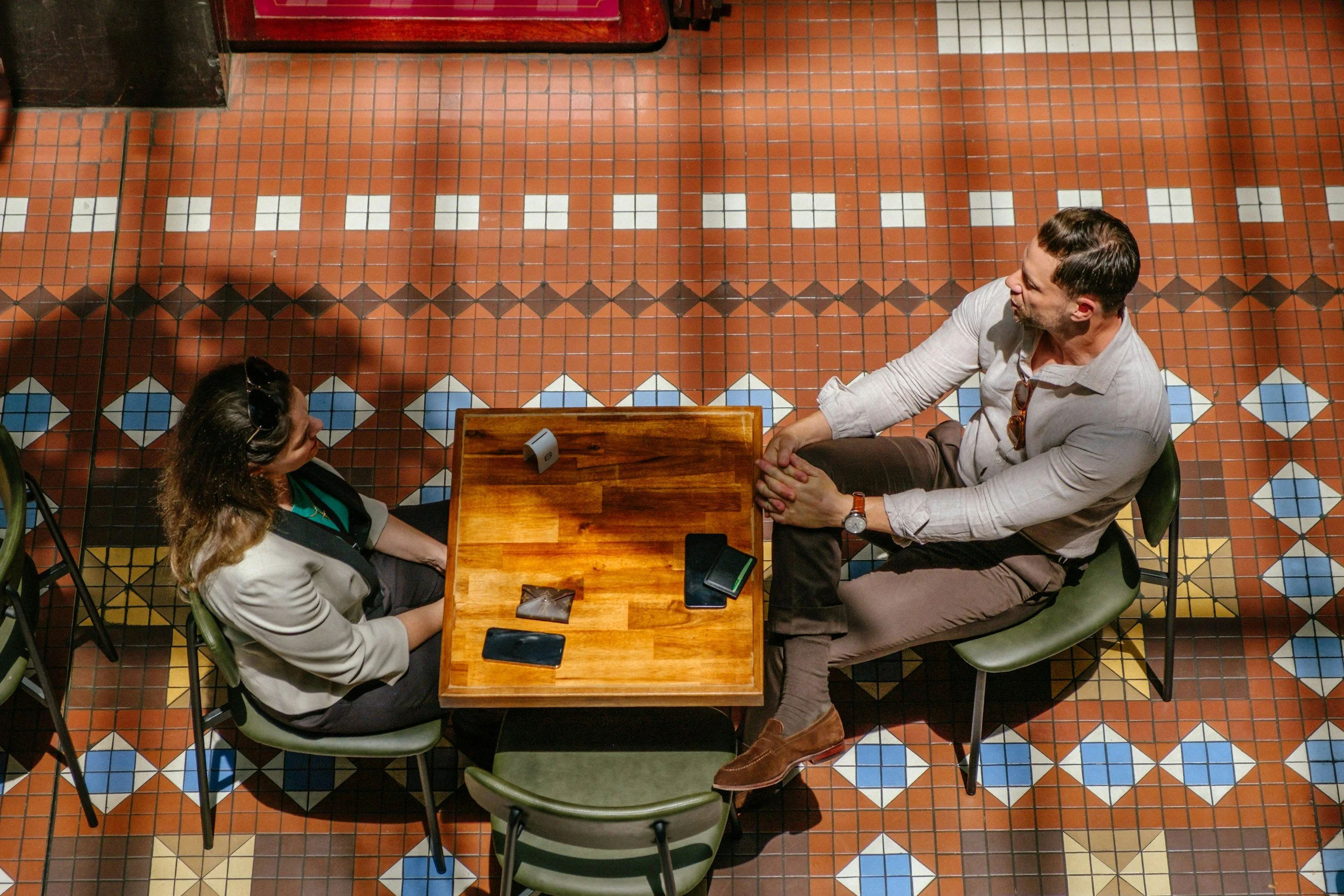 Top-down view of two people sitting across from each other at a wooden table in a restaurant, engaging in conversation. The table has two smartphones and a small white object. The floor is made of decorative tiles with geometric patterns in orange, white, blue, and black colors.