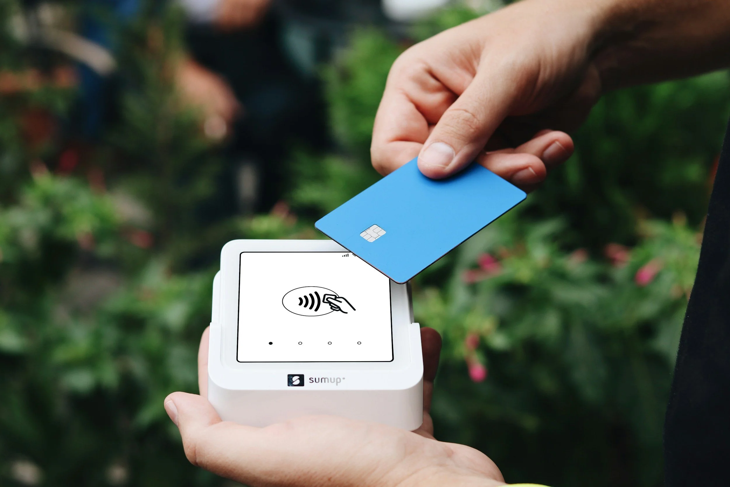 A person holding a blue credit card near a white payment device at an outdoor location with green foliage in the background.