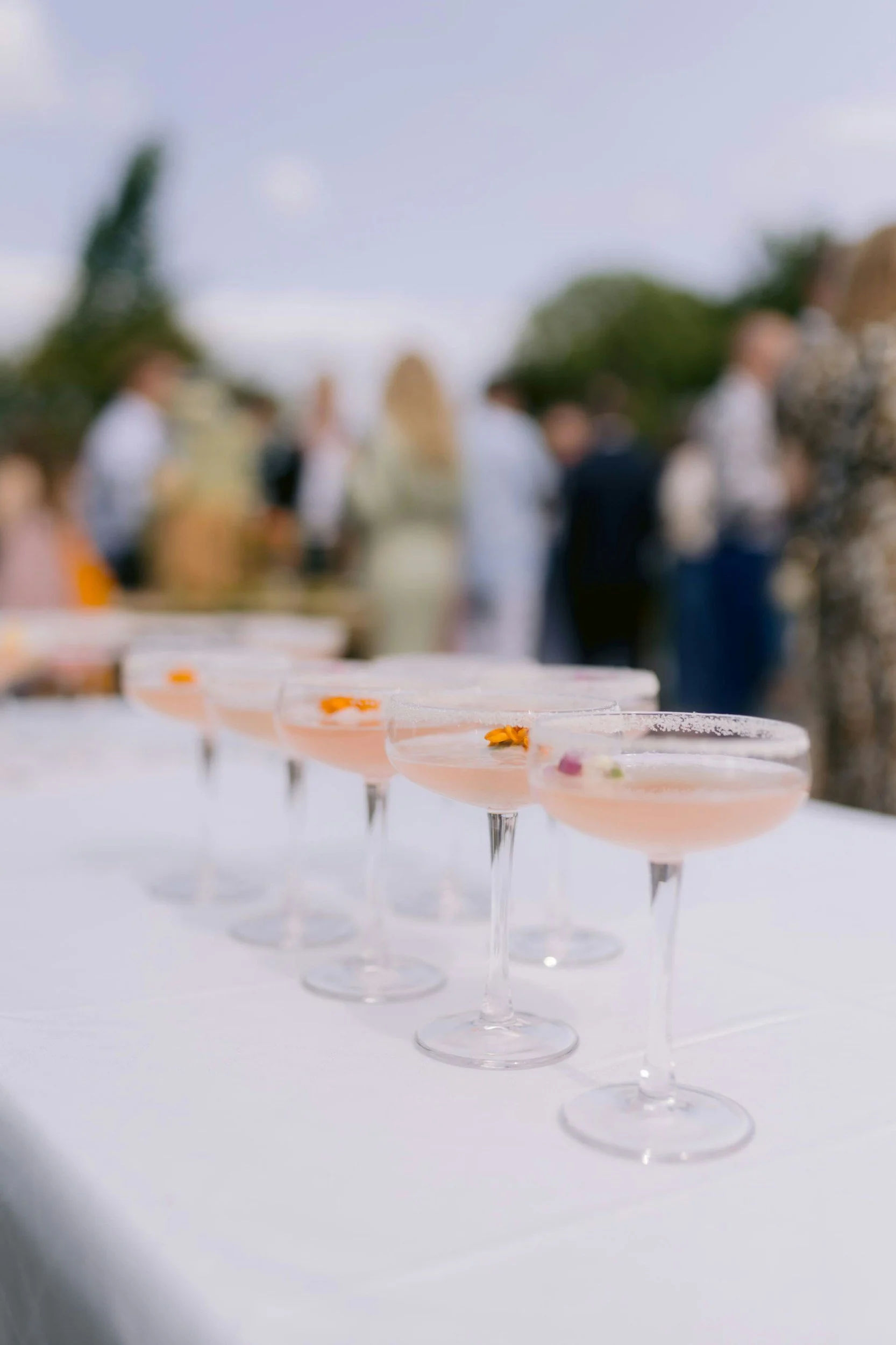 Series of pink cocktails in martini glasses with orange and purple garnishes, on a white table at an outdoor event with blurred people in the background.