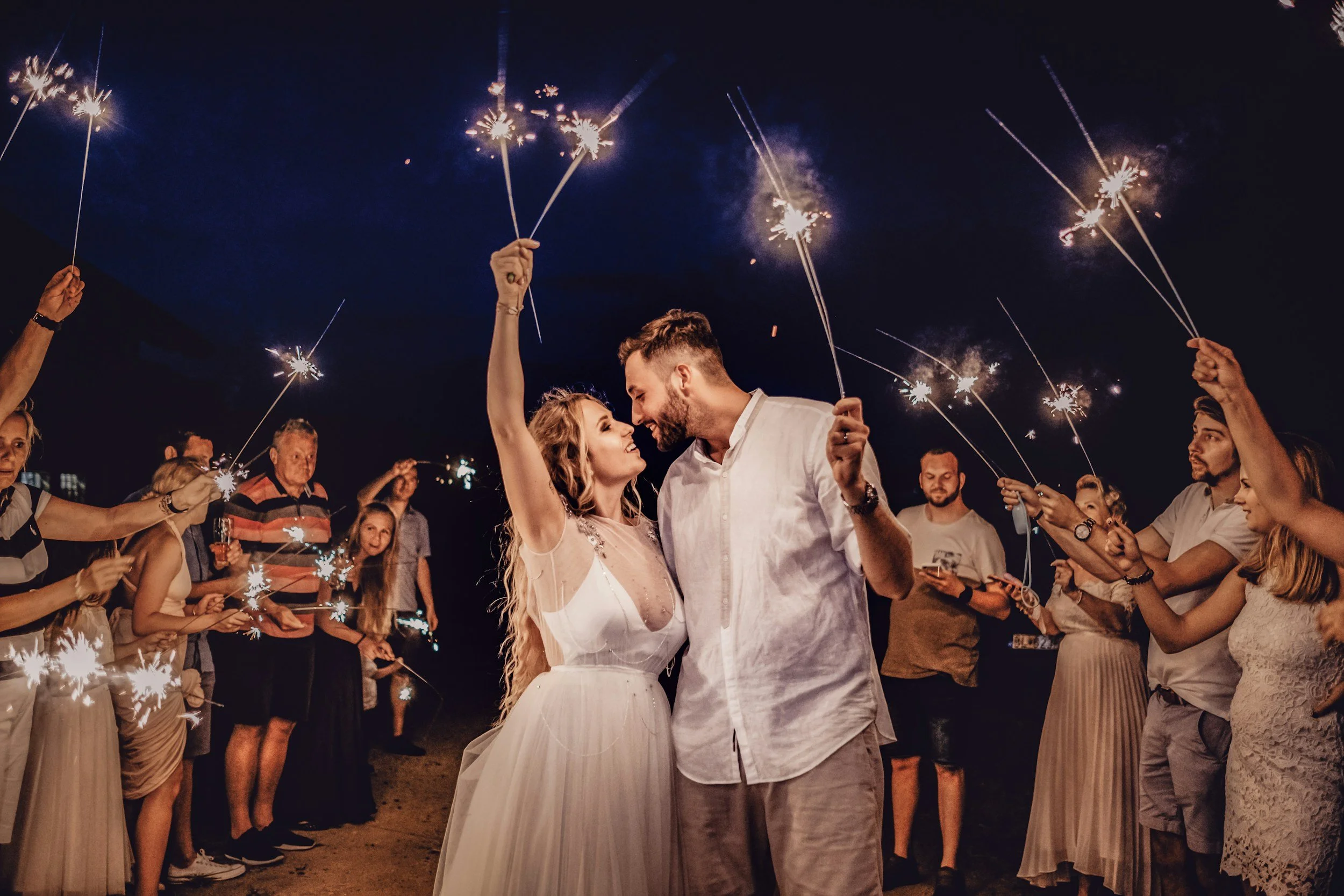 A couple in wedding attire dancing at night surrounded by friends holding sparklers.