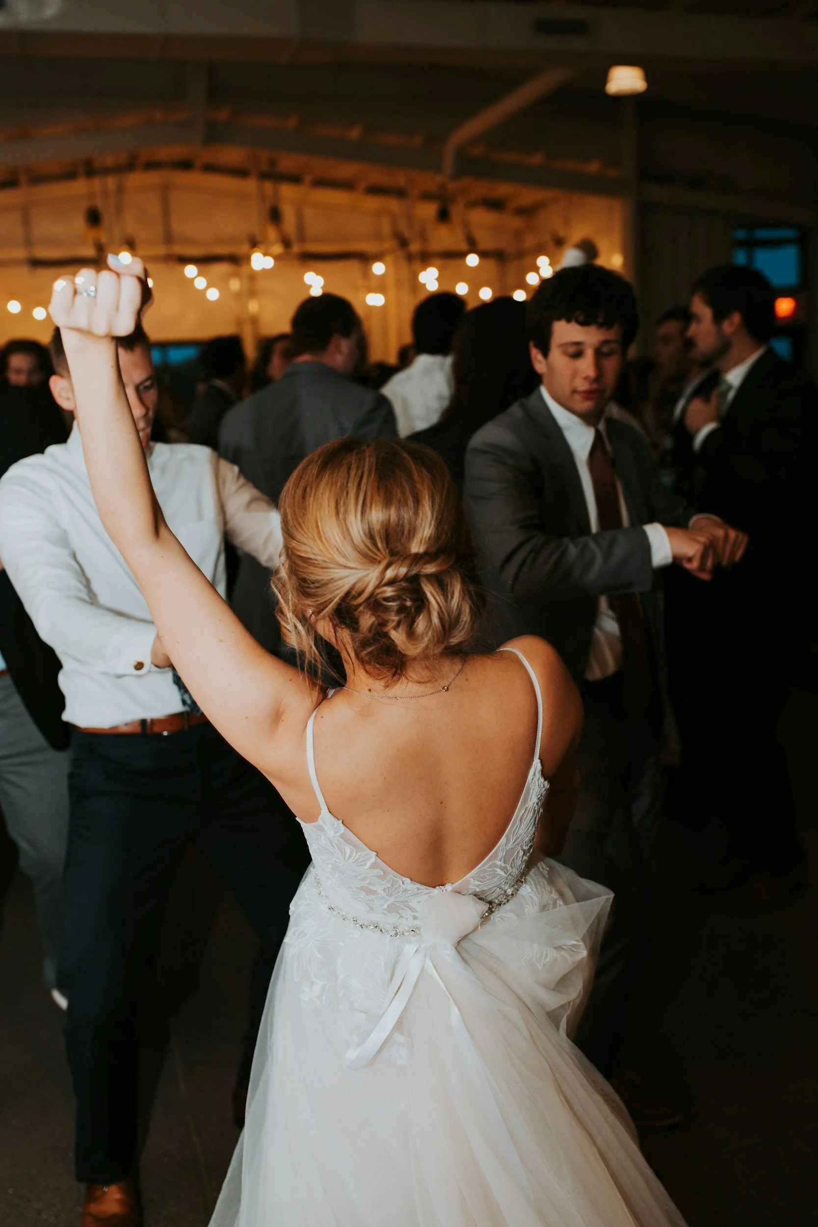 A bride with styled hair dancing in a white wedding dress, with a young man in a gray suit and other guests dancing and socializing at a party in a warmly lit venue.