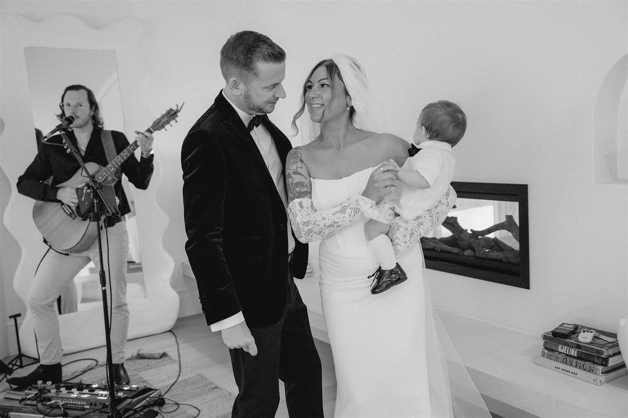 A newlywed couple, a man in a tuxedo and a woman in a lace wedding dress, are standing together, smiling, and looking into each other's eyes while holding a toddler child dressed in white with a bow tie. A musician with long hair, glasses, and an earpiece is playing an acoustic guitar and singing into a microphone in the background. There is a stack of books and a remote control on a white table near a fireplace in a minimalistic, modern room.