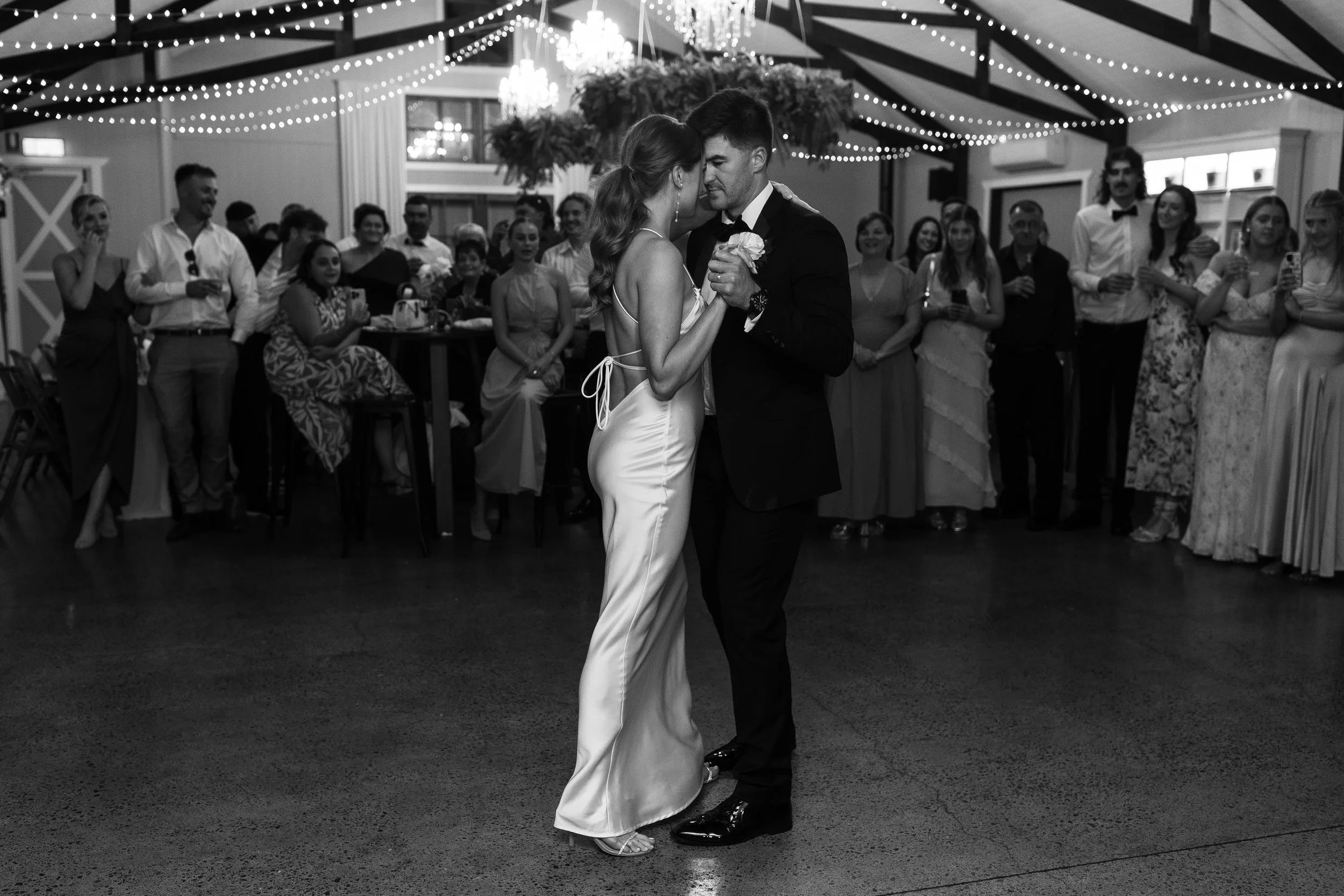 A couple dancing closely at a wedding reception with guests watching and smiling, decorated with string lights and chandeliers.