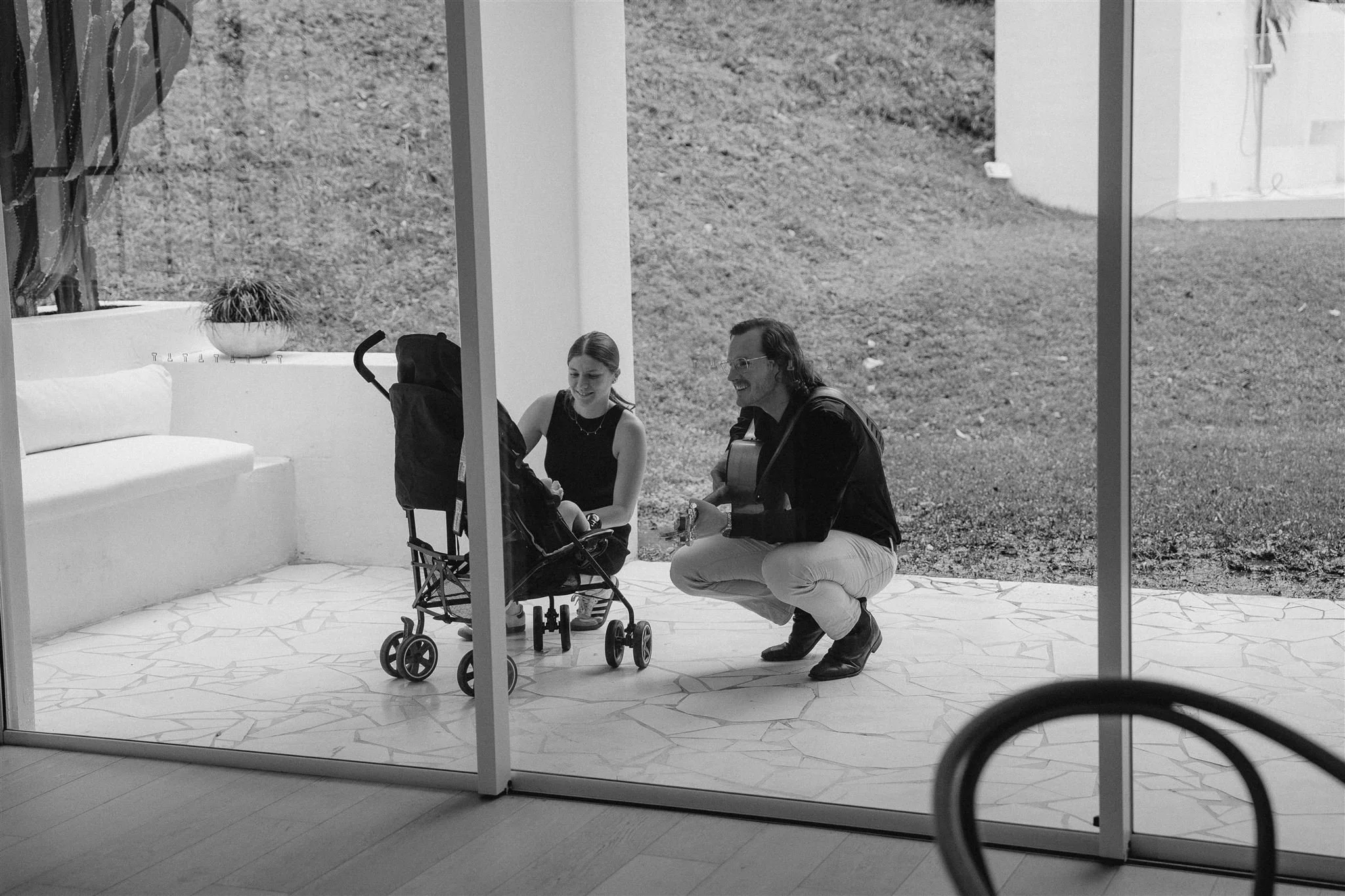 Two people, a woman and a man, are sitting and kneeling on a tiled patio outside a building with large windows. The woman has a stroller nearby, and they are smiling and engaging with each other, with the man holding a guitar. There is a white bench with a potted plant on it, and some grass and a building are visible outside.