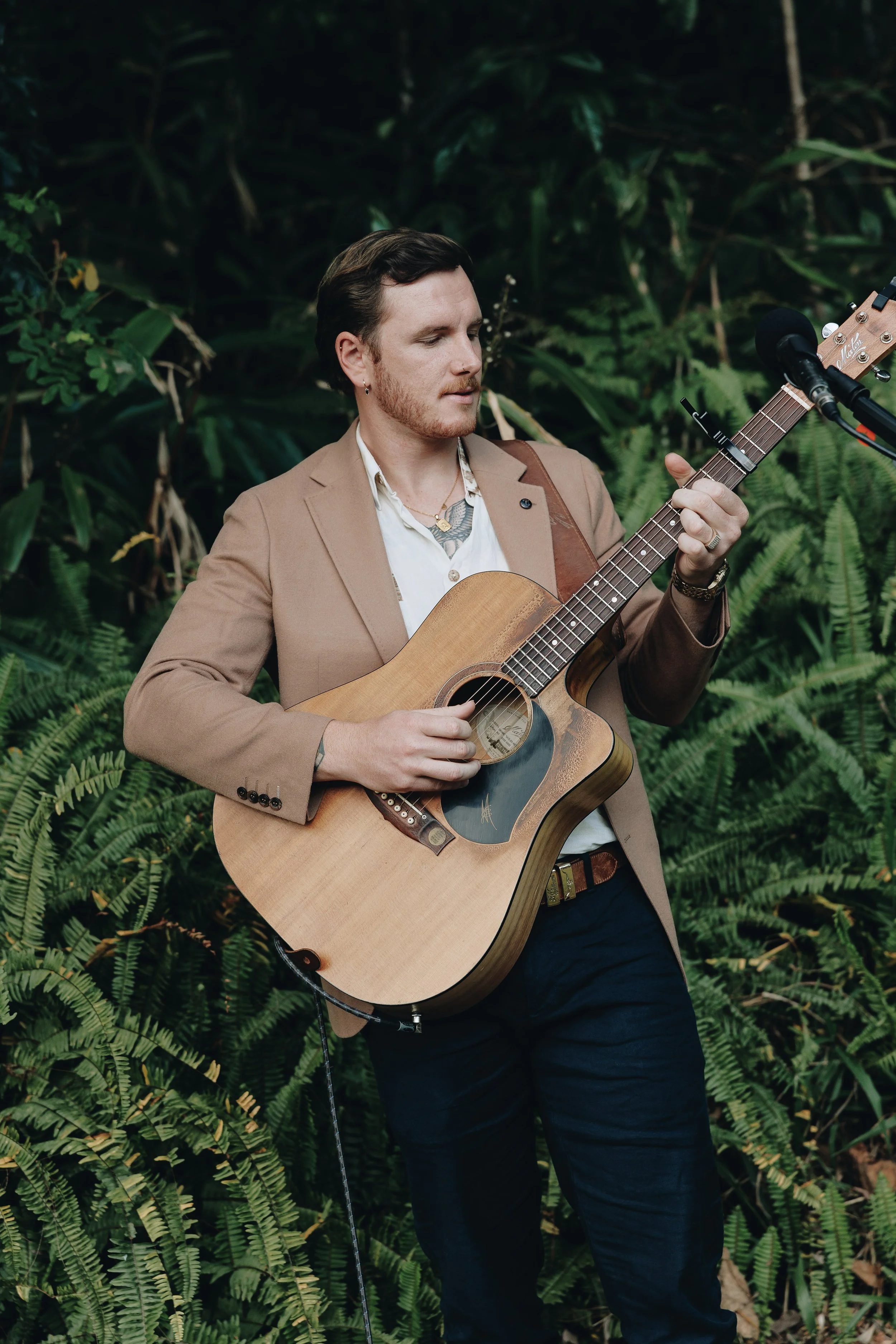 A man with dark hair and beard playing an acoustic guitar outdoors among lush green foliage.