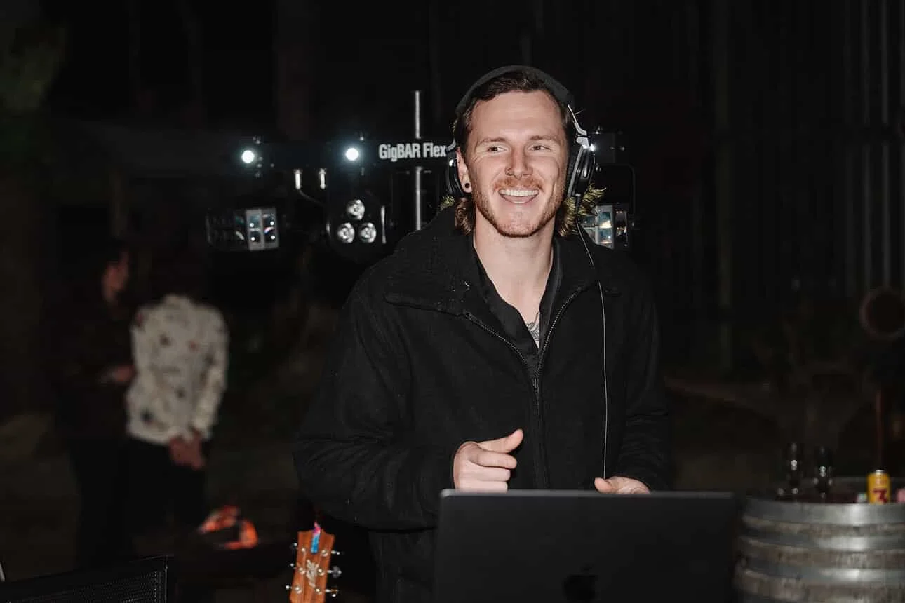A smiling man wearing headphones stands behind a DJ setup in a dark, outdoor venue, with people in the background.