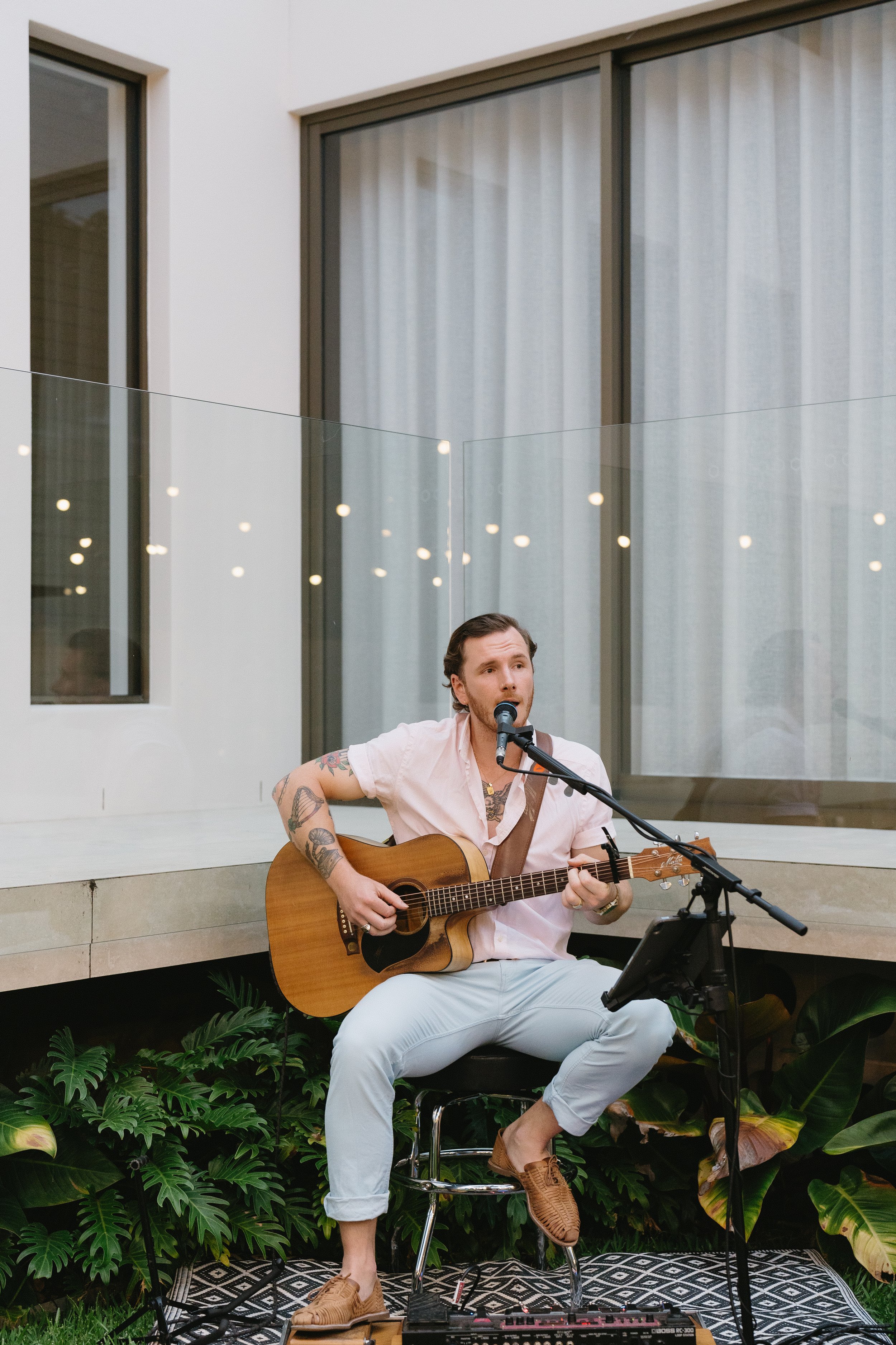 A man with tattoos on his arms is sitting outdoors on a stool, playing an acoustic guitar and singing into a microphone. He is wearing a light pink shirt, white pants, and brown shoes. There are plants behind him and large windows with curtains in the background. Music equipment and a tablet are in front of him.