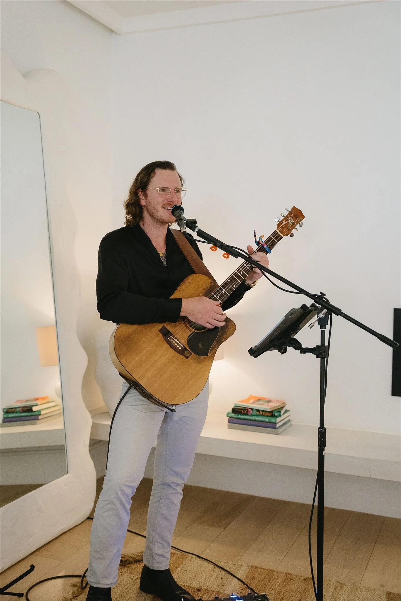 A man with long brown hair, glasses, wearing a black shirt and white pants, playing an acoustic guitar and singing into a microphone in a room with white walls and books on the shelves.
