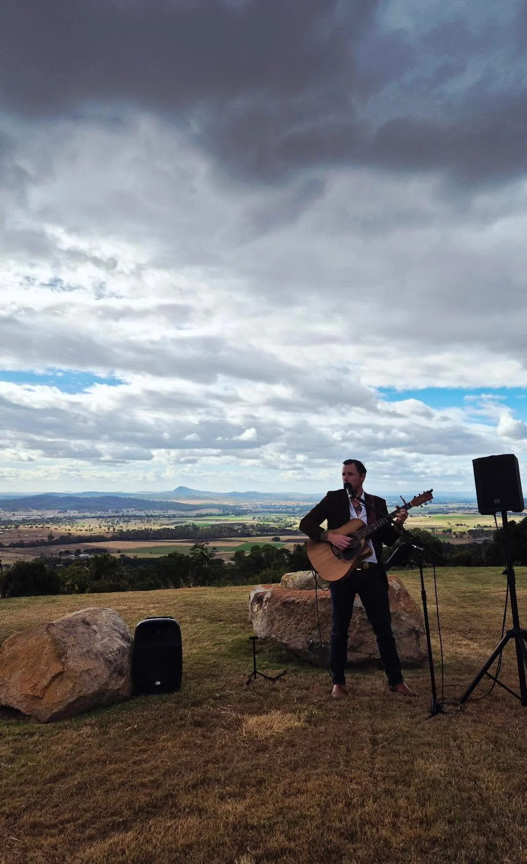 A man dressed in a blazer and dress shirt playing an acoustic guitar outdoors on a grassy hill, with large rocks, a scenic landscape, and a dramatic cloudy sky in the background.