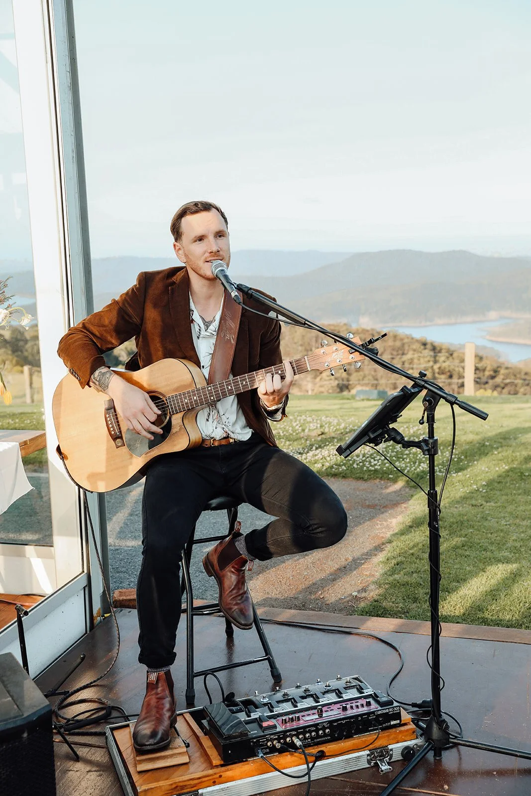 A man playing an acoustic guitar and singing into a microphone outdoors with a scenic landscape of mountains, a lake, and a grassy area in the background.