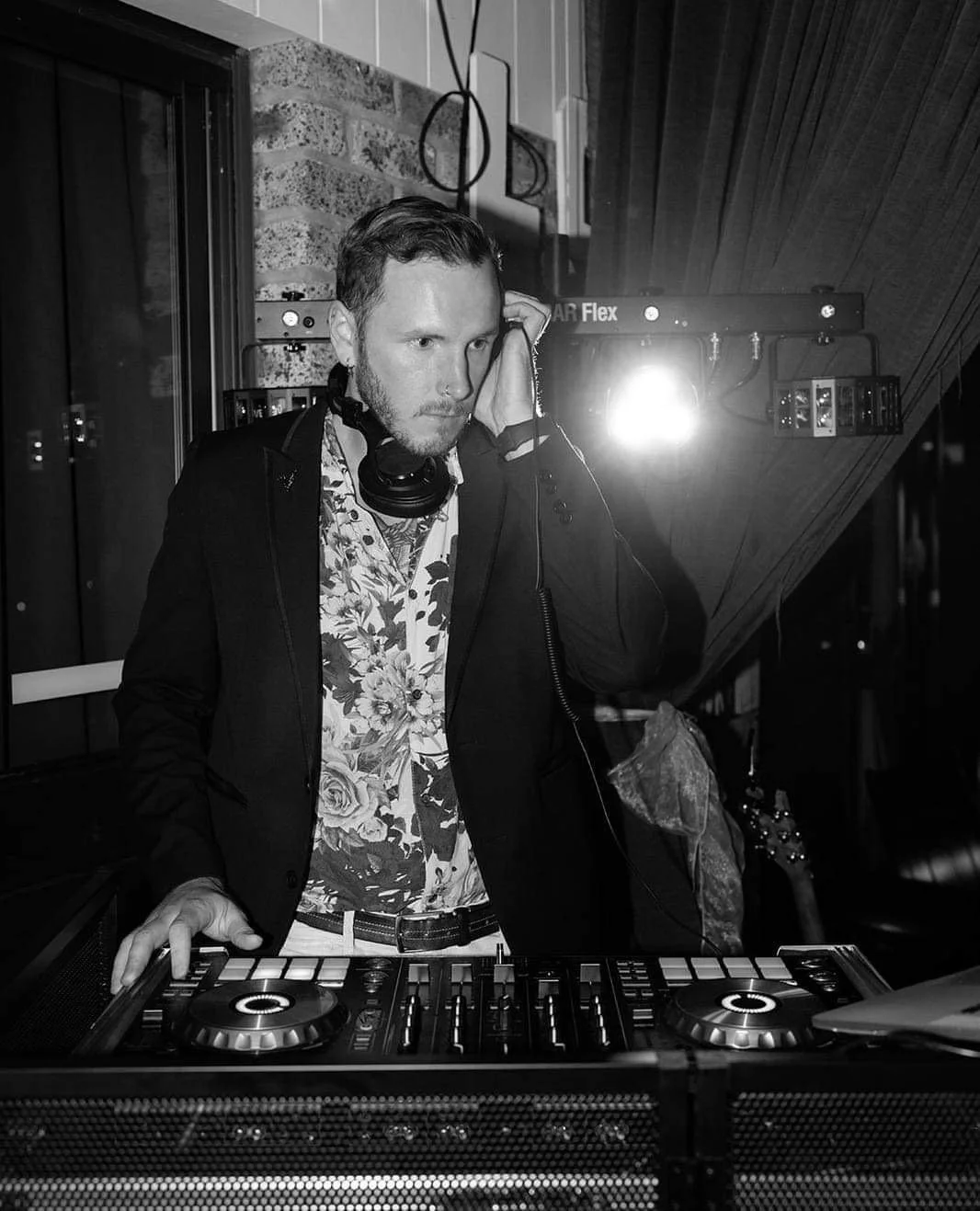 A man DJing at a party, wearing a floral shirt, black jacket, and headphones around his neck, with turntables in front of him, in a dimly lit venue with brick and wood walls.