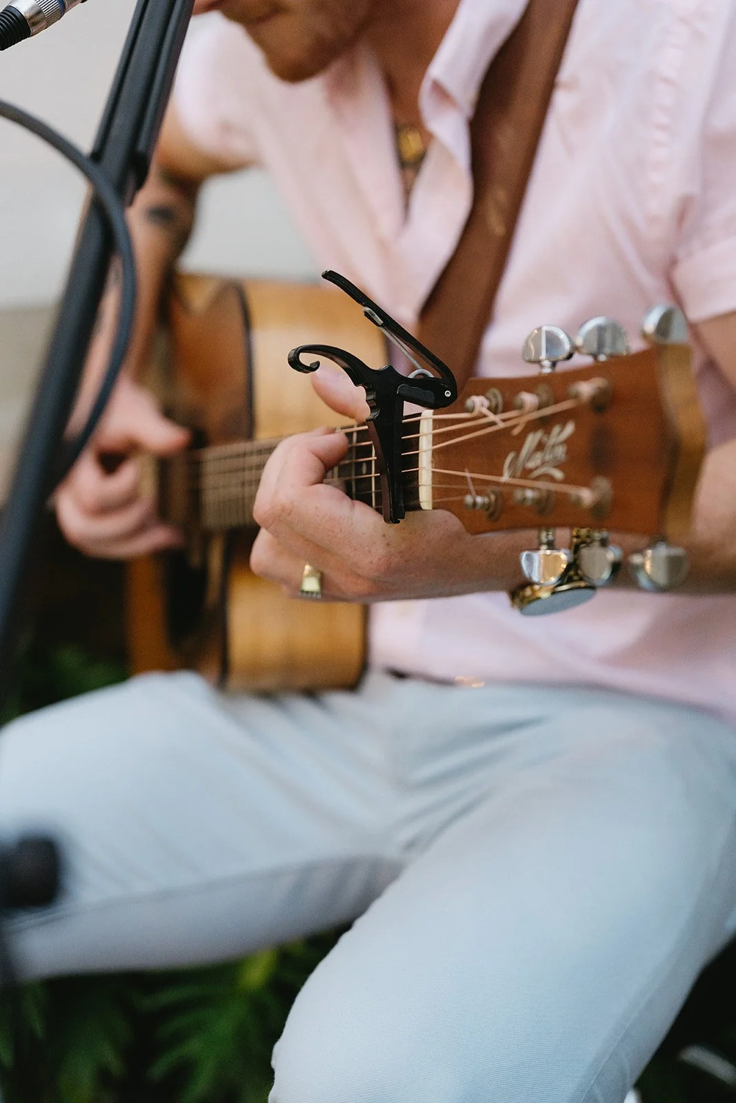 Person playing acoustic guitar with a capo on the neck, wearing a pink shirt and white pants, sitting outdoors.